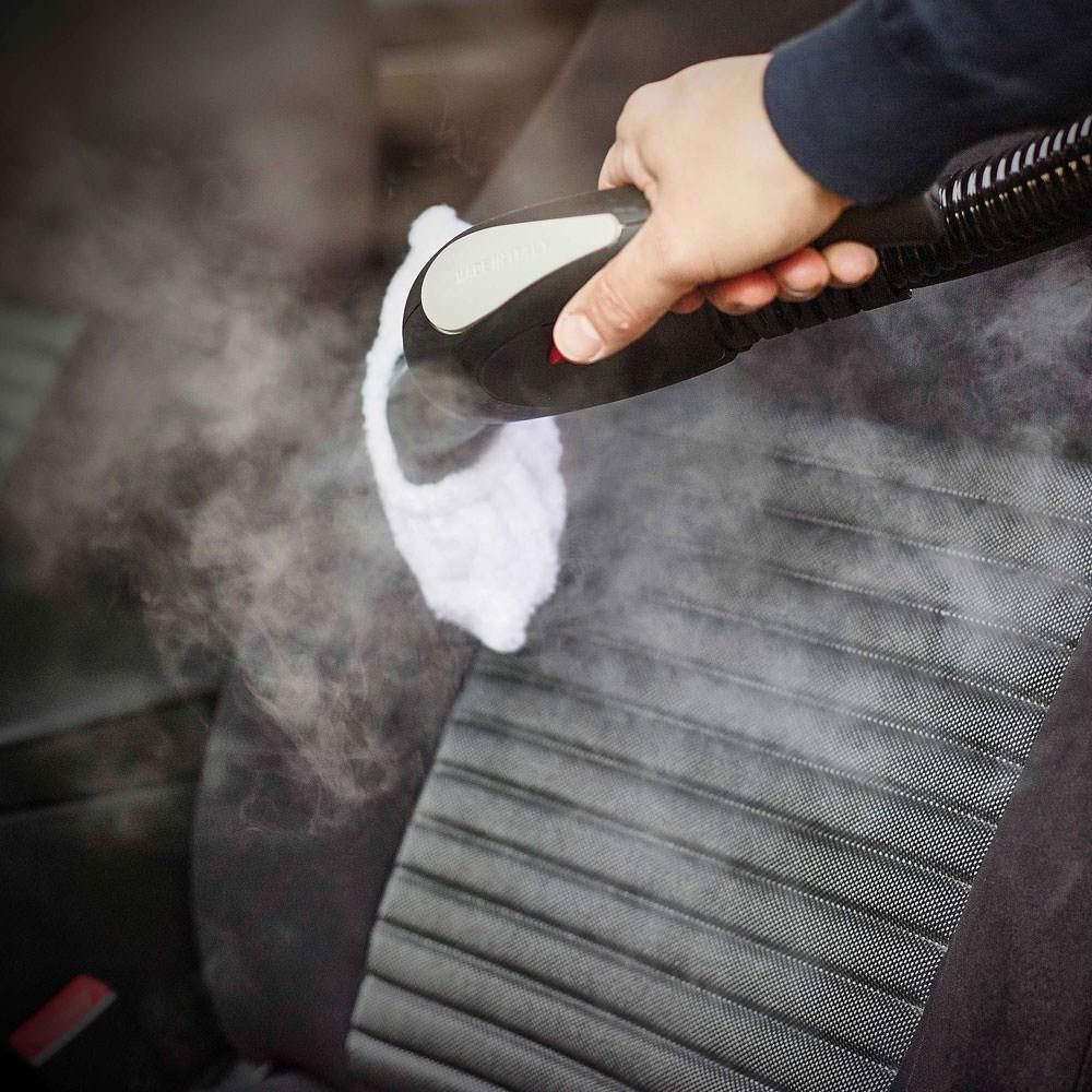 Ręka parująca fotel samochodowy za pomocą parownicy, wytwarzając parę. Siedzisko jest szare i teksturowane.