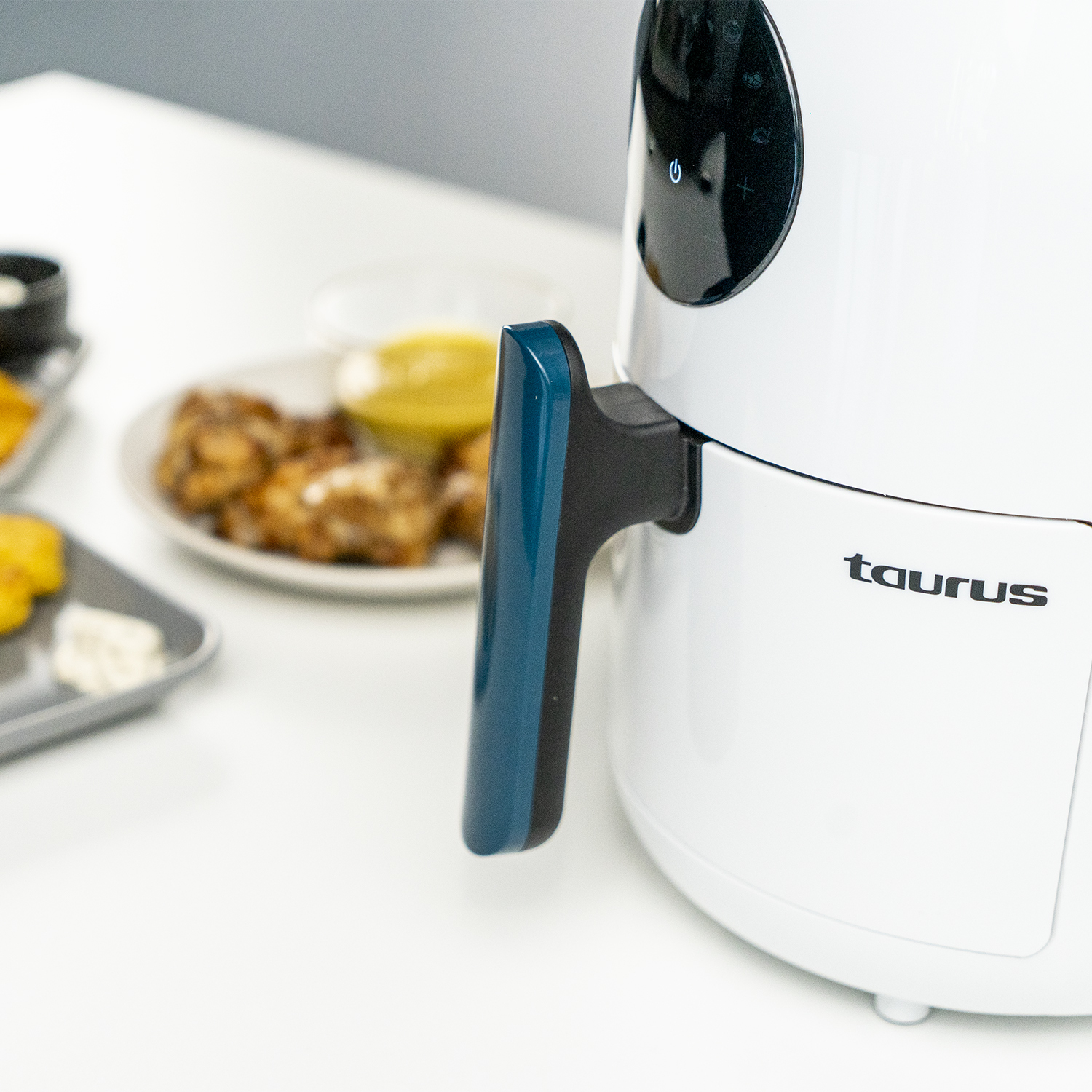 Freidora de aire blanca con asa azul. Comida en plato en el fondo. Marca: Taurus.