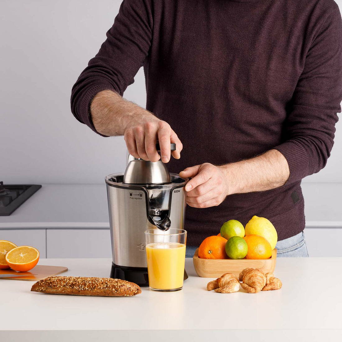 Mężczyzna używa wyciskarki do soku z pomarańczami, cytrynami, chlebem i rogalikami na białym blacie.