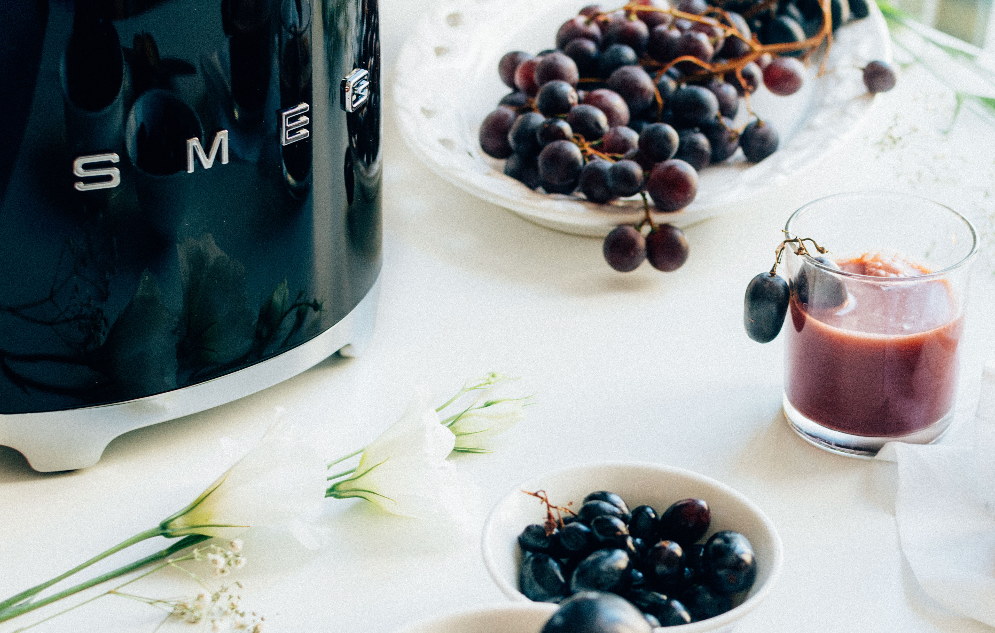 Tostadora SMEG negra, uvas y un vaso con una bebida morada sobre una mesa blanca.