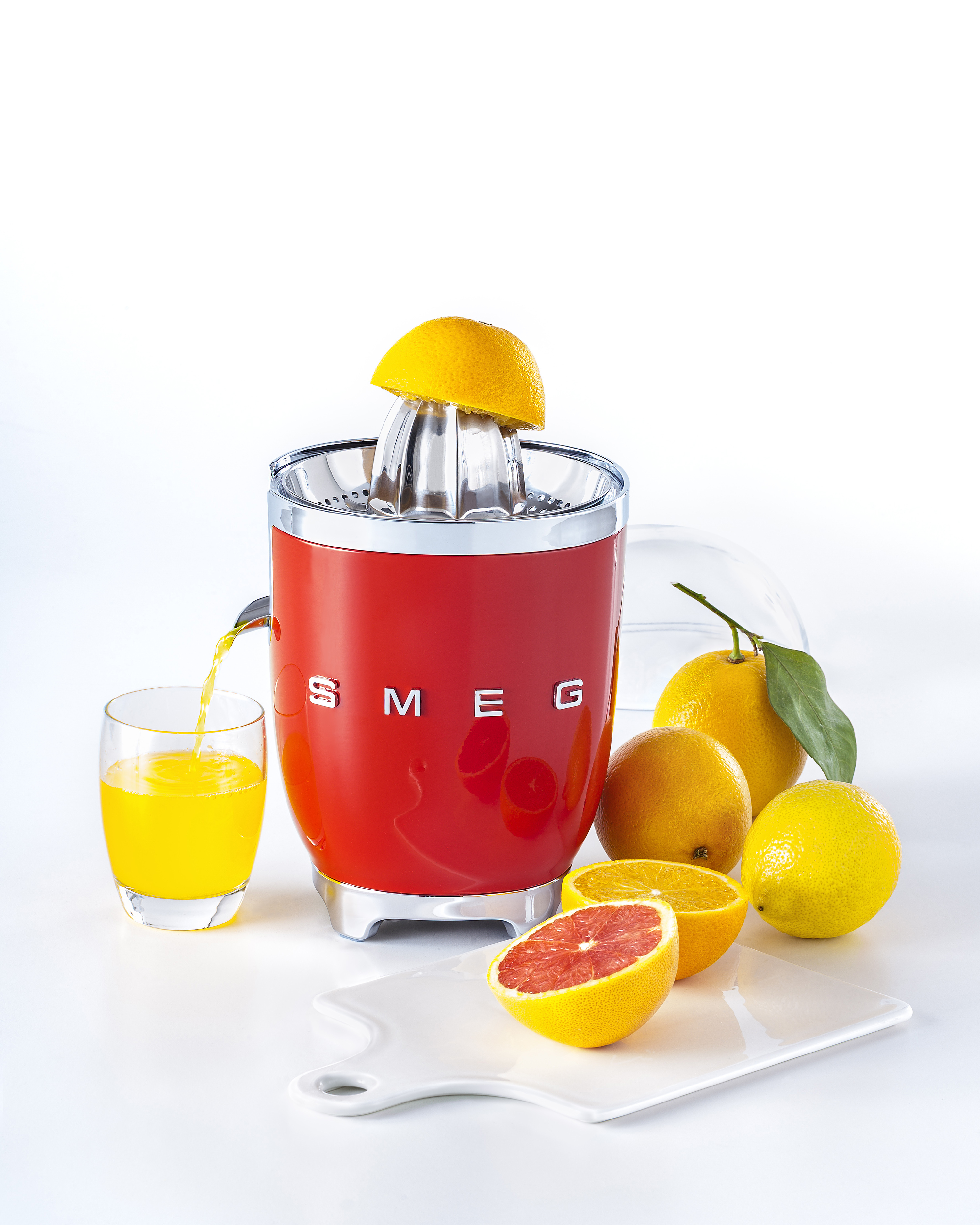 Roter Smeg Entsafter mit Orangen, Saft im Glas und Schneidebrett mit halber Orange.