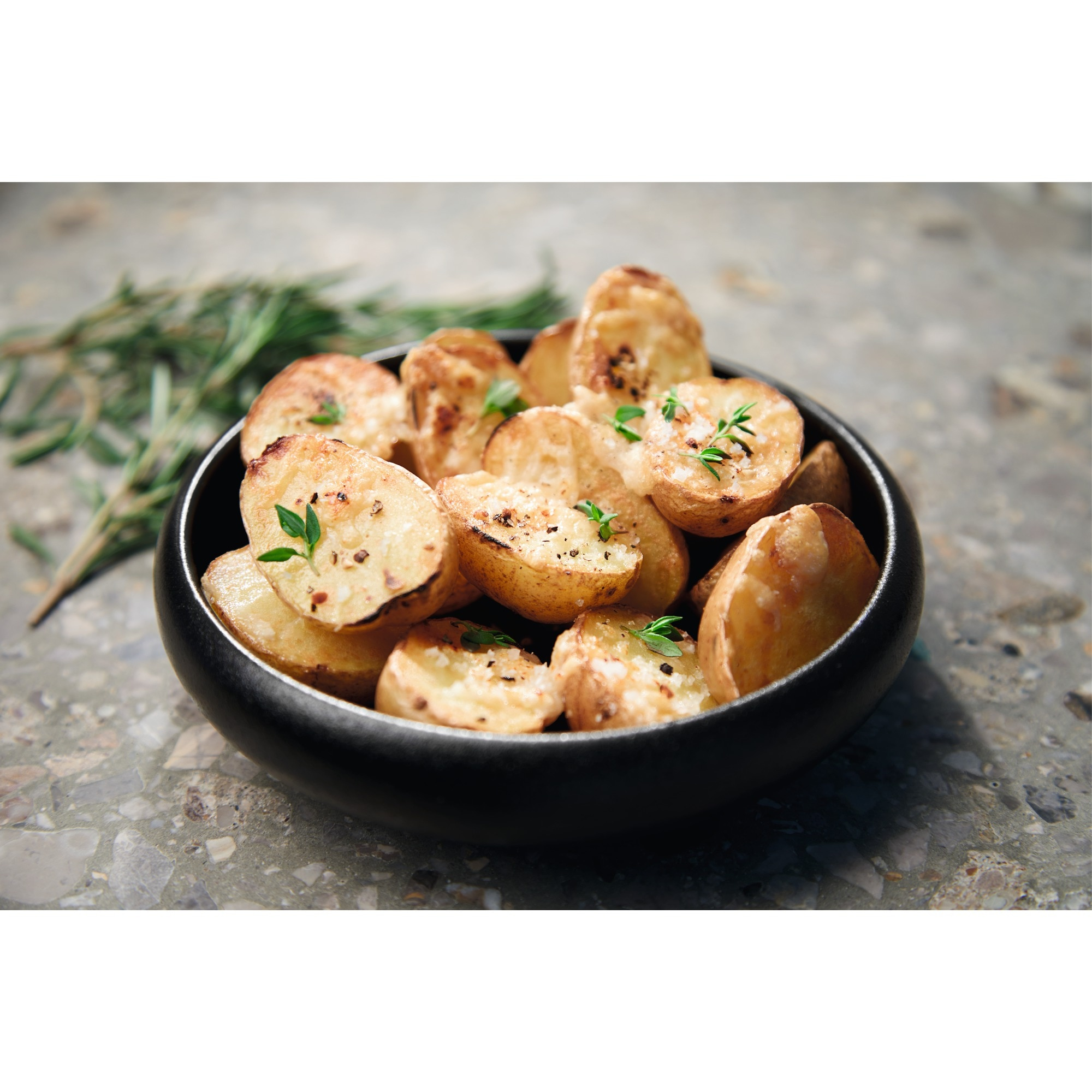 Czarna miska wypełniona pieczonymi ziemniakami. Świeże gałązki rozmarynu leżą obok miski.