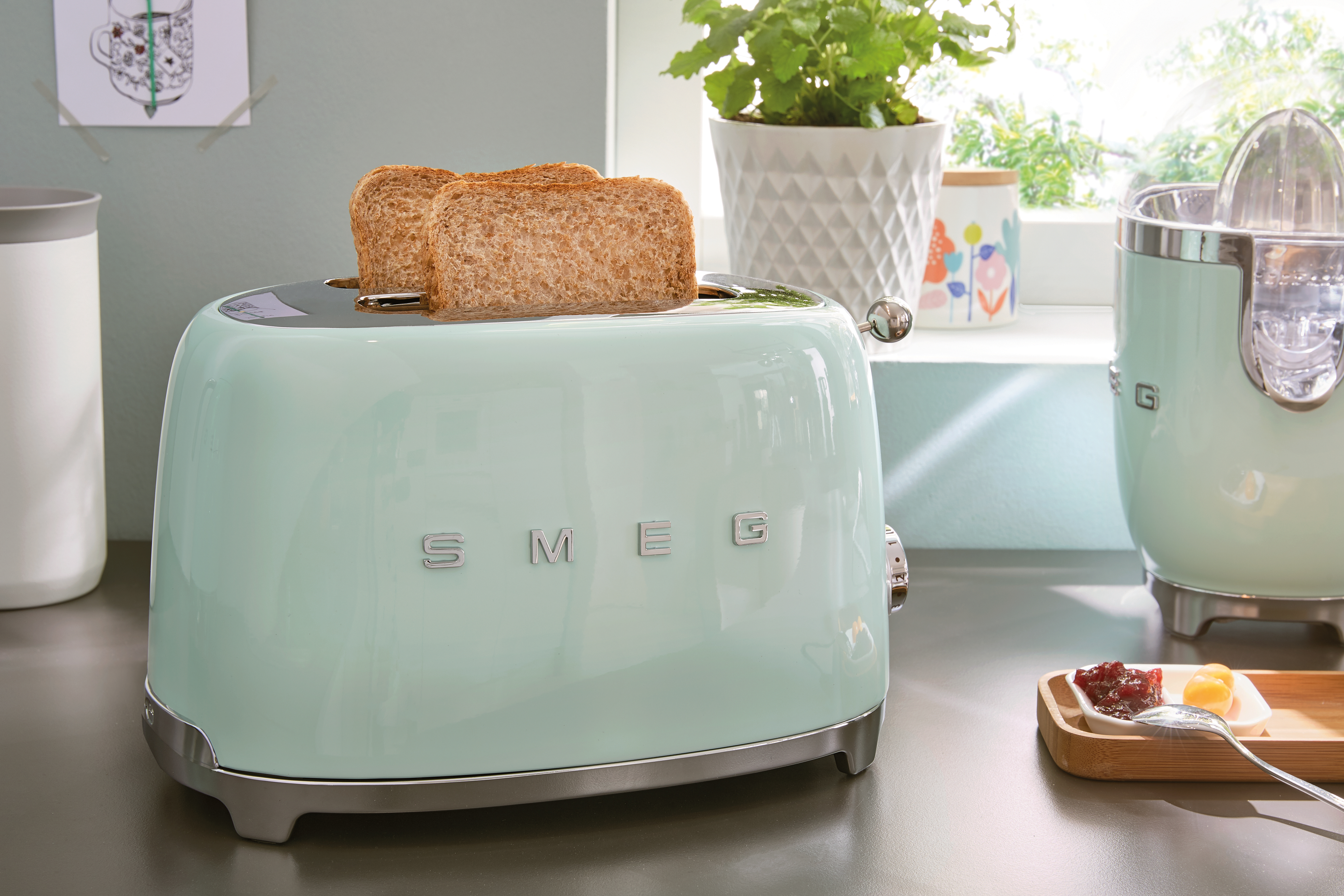 Un tostapane SMEG verde menta con pane tostato. Altri oggetti da cucina sono visibili sullo sfondo.