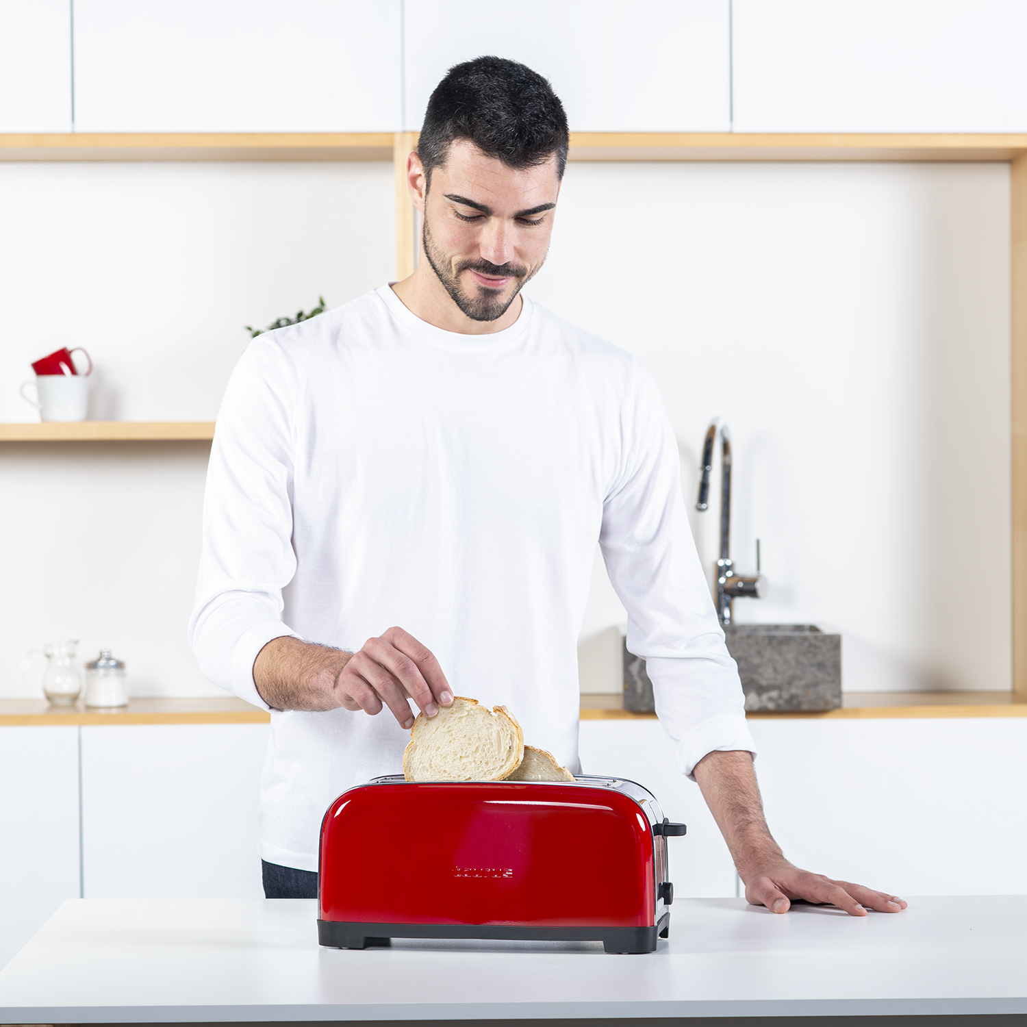 Mężczyzna wkłada kromki chleba do czerwonego tostera, stojąc przy białym blacie.