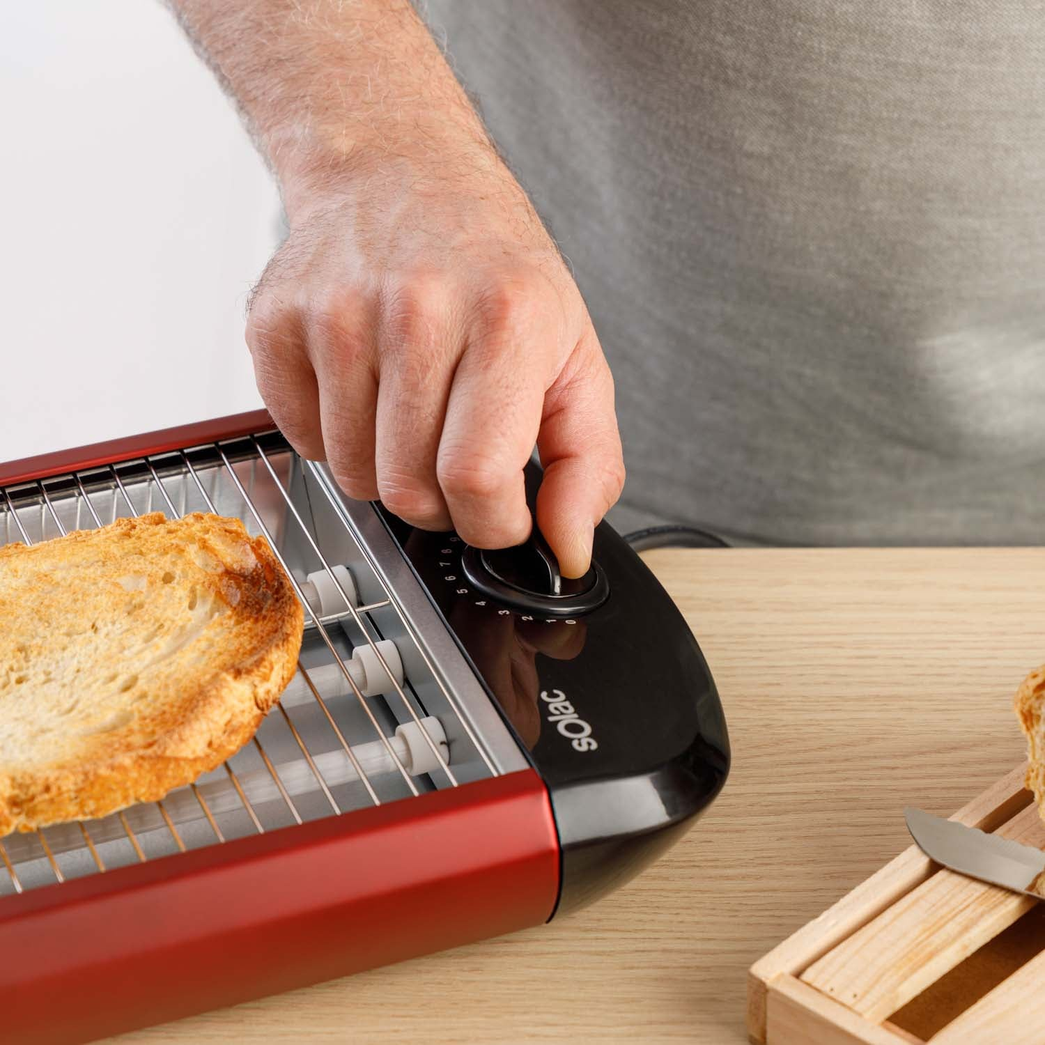 Ręka osoby regulująca pokrętło na czerwonym tosterze z tostami w środku. Drewniany stół.