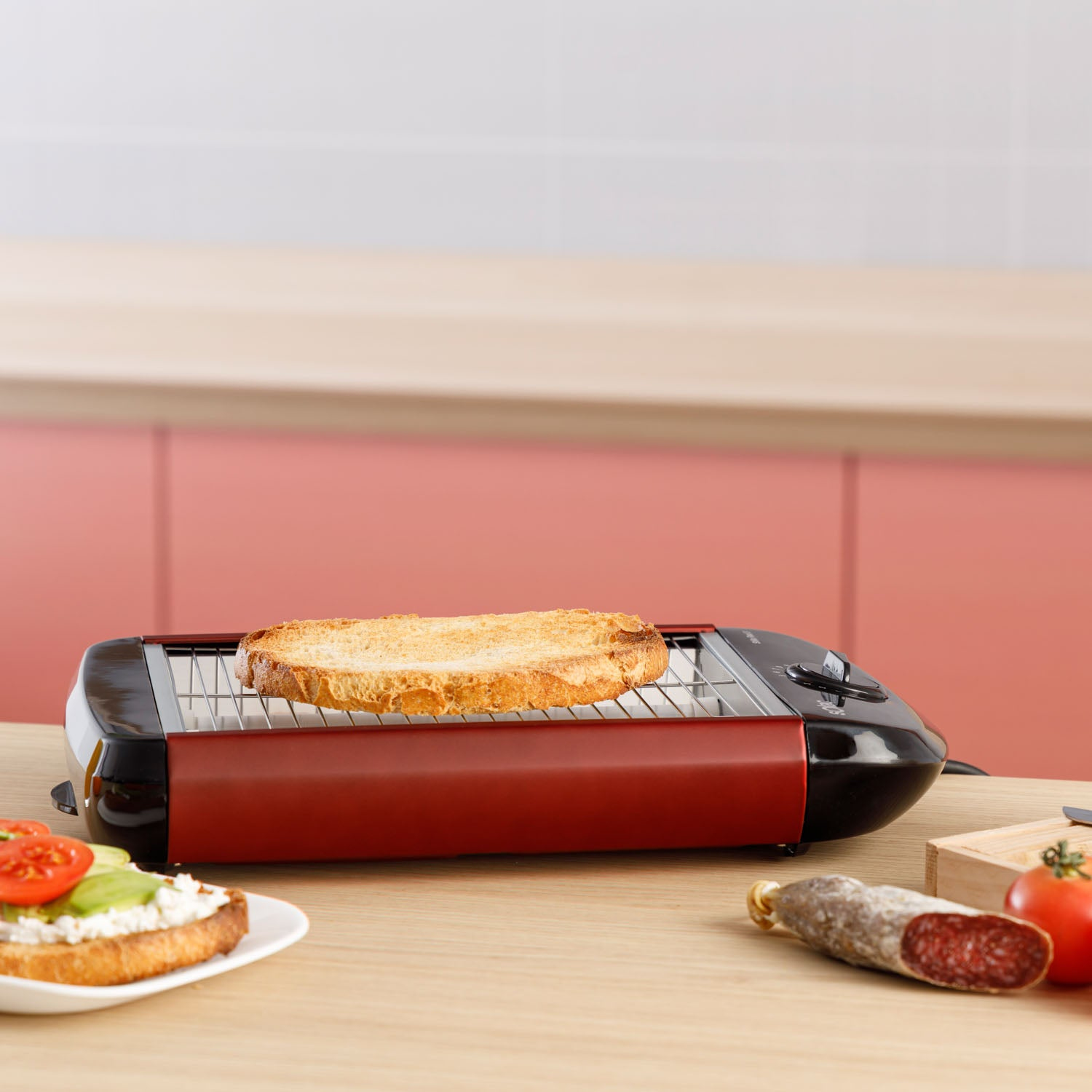 Tost na czerwonym grillu, kanapka, kiełbasa, pomidor. Drewniany stół, różowa ściana.