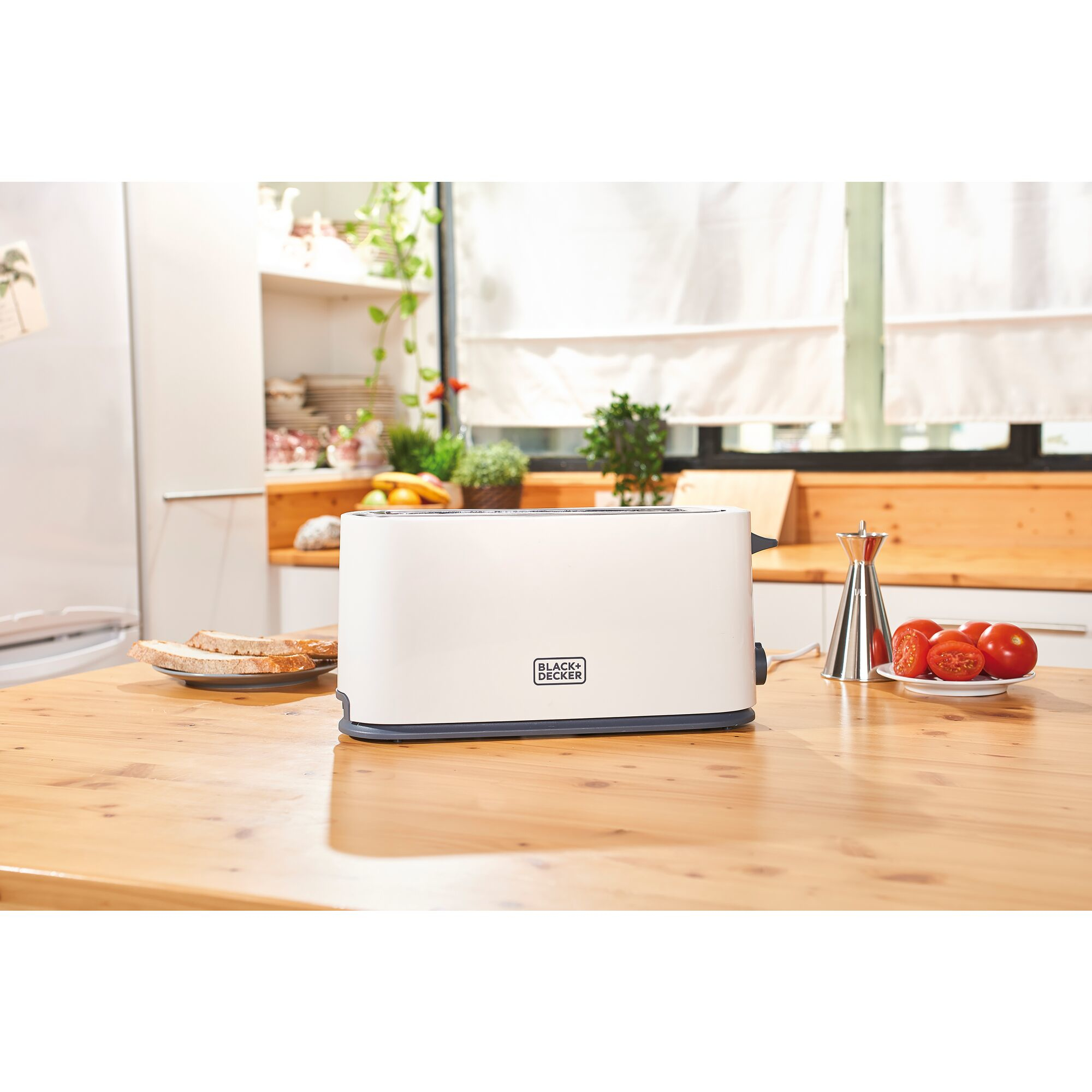 Biały toster na drewnianym stole z chlebem, pomidorami i oknem w tle.