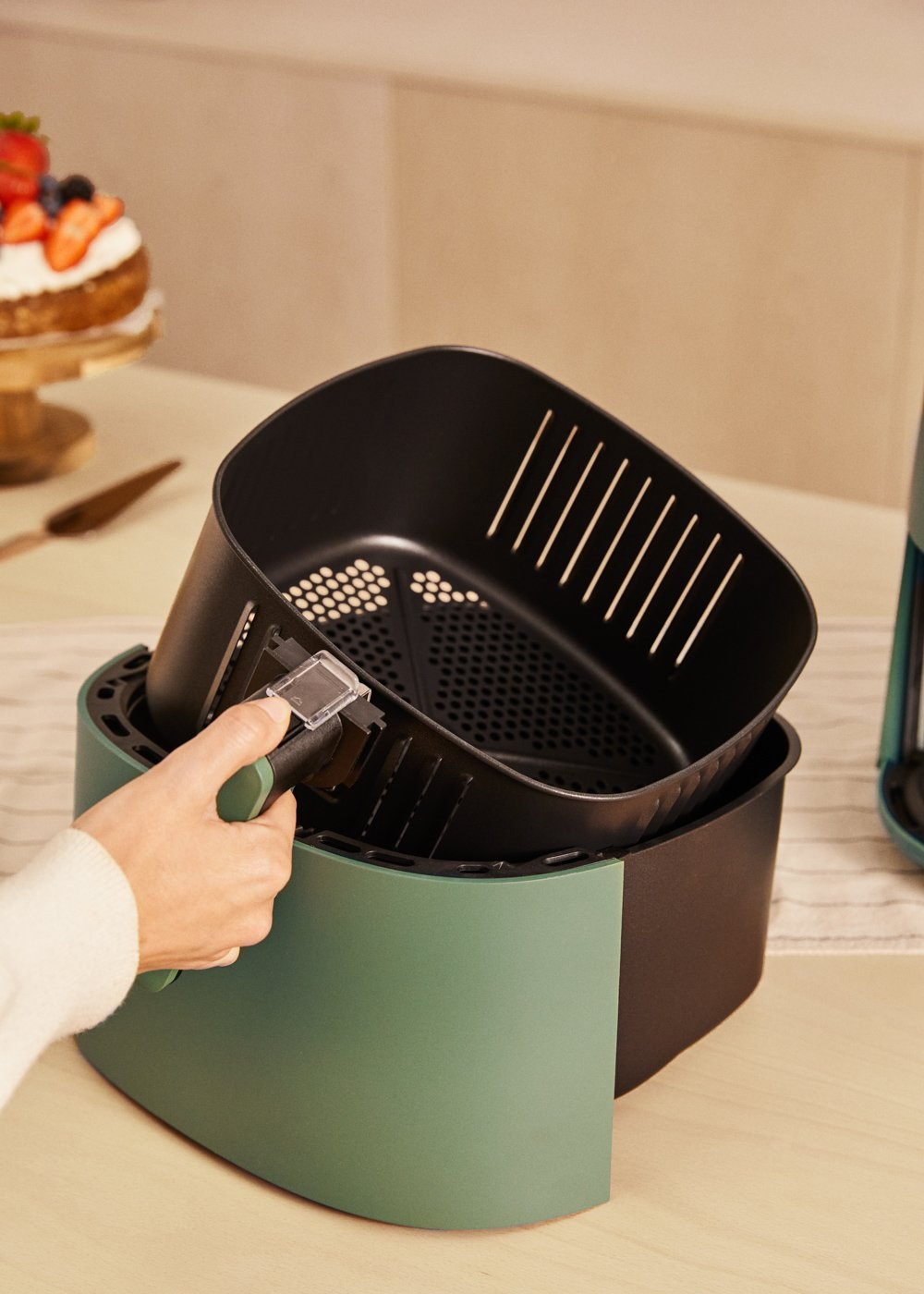 Zielona i czarna frytkownica powietrzna z podnoszonym koszem. Dłoń kobiety podnosi kosz.