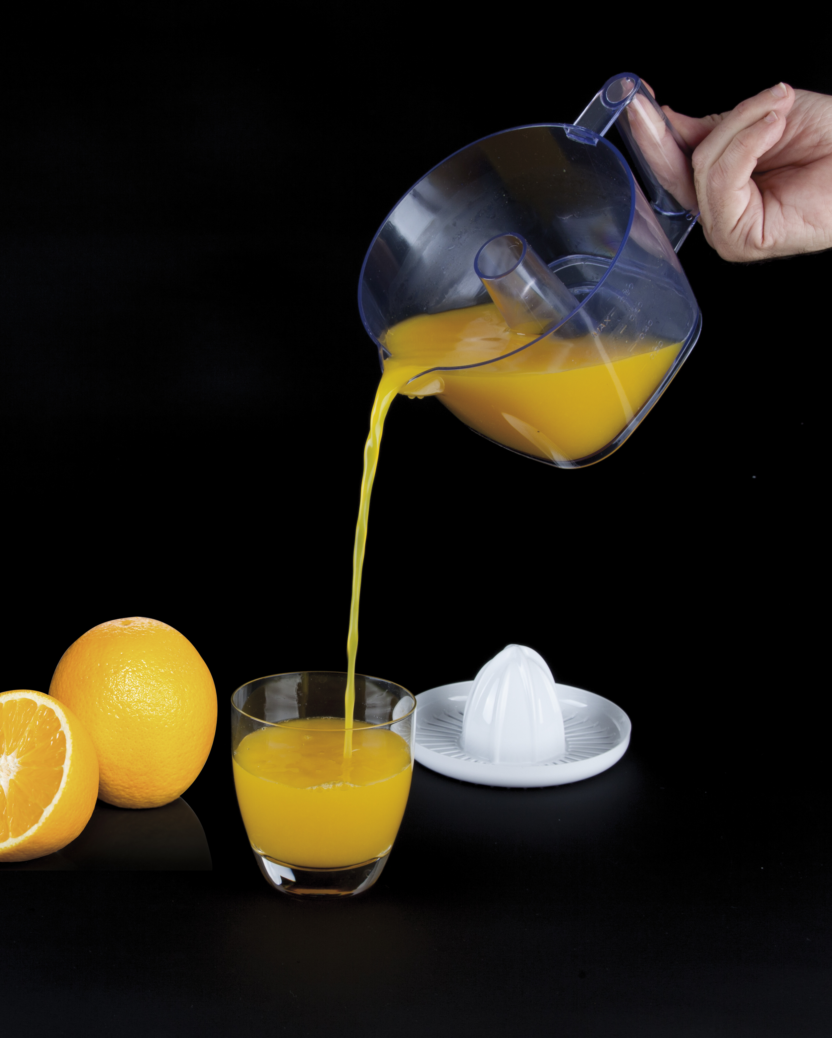 Jugo de naranja vertiéndose de una jarra a un vaso. Naranjas y exprimidor sobre una superficie negra.