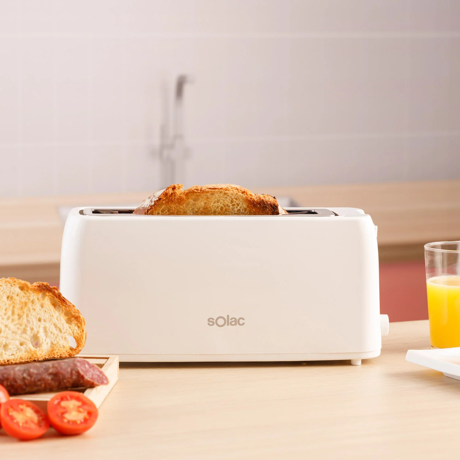 Biały toster z dwoma kromkami tostowanego chleba. Drewniana deska z kiełbasą i pomidorami.