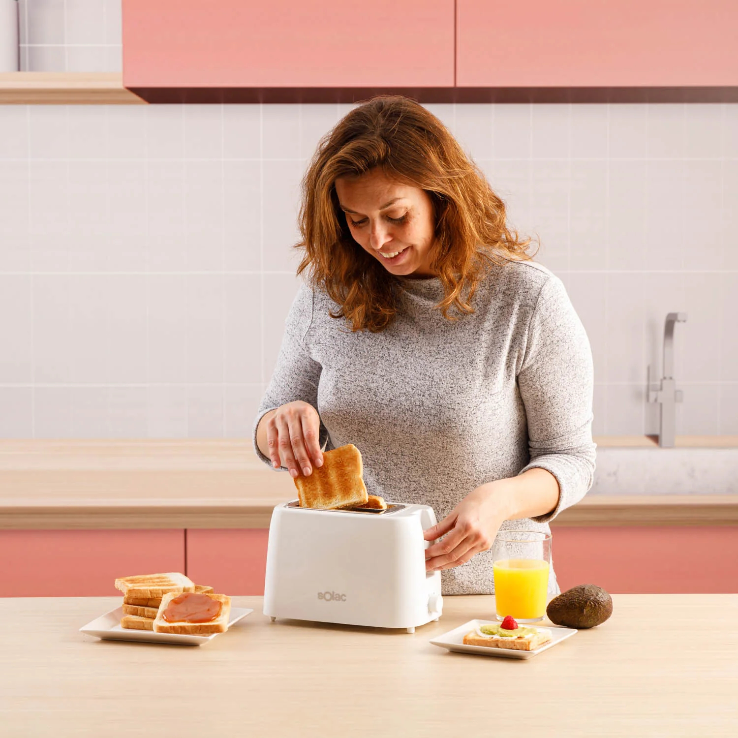 Kobieta wkłada tosty do białego tostera. Na stole są inne talerze z tostami, sok pomarańczowy i awokado.