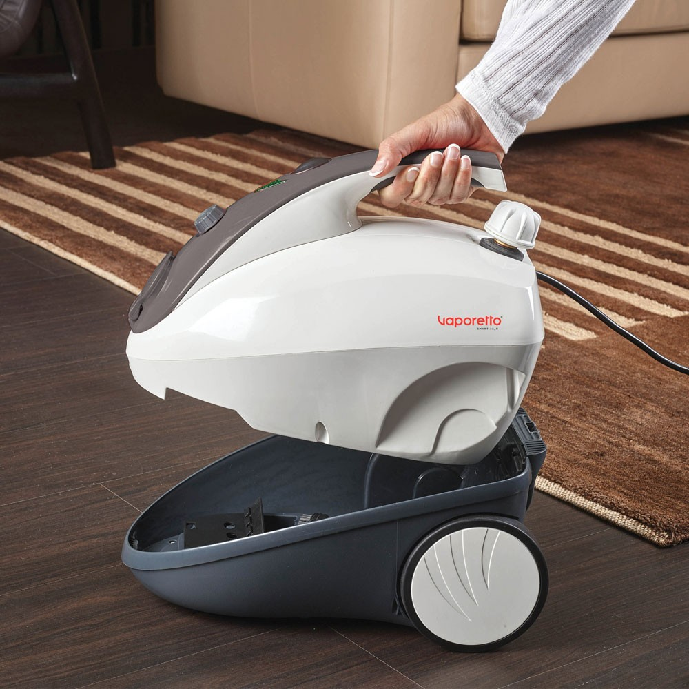 Ręka osoby trzymająca biało-szarą myjkę parową. Brązowy dywan.