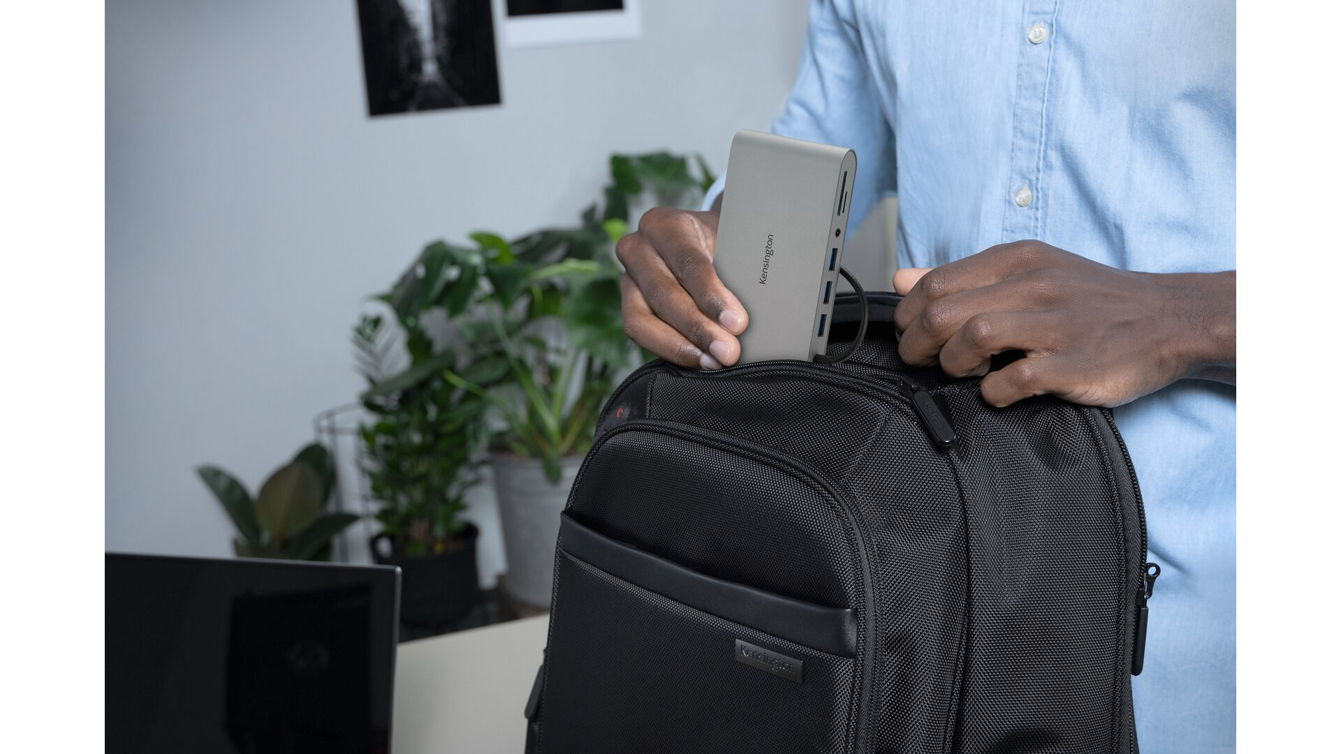 Eine Person legt ein graues Tech-Gerät in einen schwarzen Rucksack, im Hintergrund eine Pflanze und ein Schreibtisch.