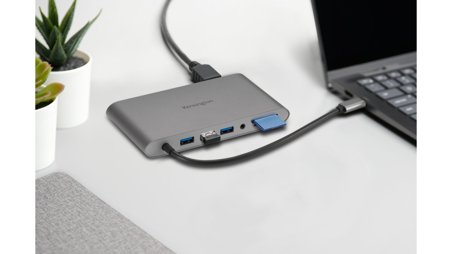 Silberne Kensington USB-C Dockingstation, an einen Laptop angeschlossen. Auf einem weißen Schreibtisch.