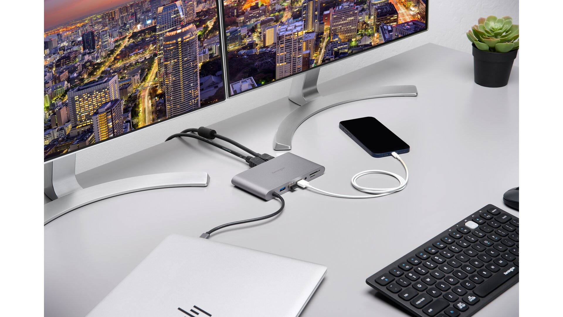Schreibtisch mit zwei Monitoren, Laptop, Telefon und Hub. Stadtlandschaft im Hintergrund. Schwarze Tastatur und Pflanze.