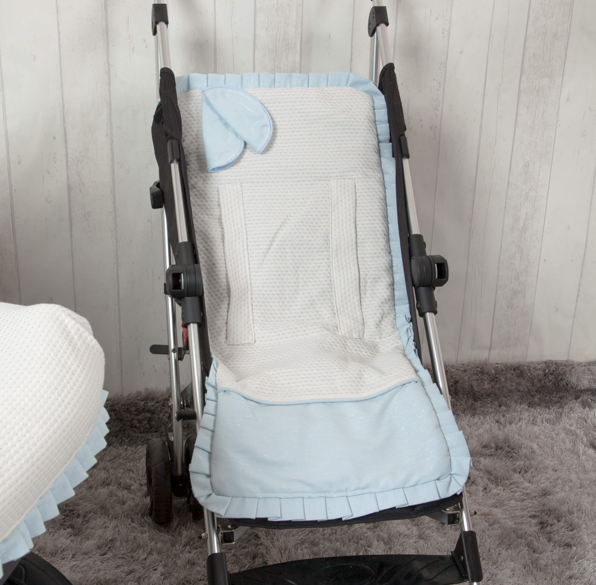 Asiento de cochecito para bebé con detalles azules y volantes. Alfombra gris. Fondo blanco.