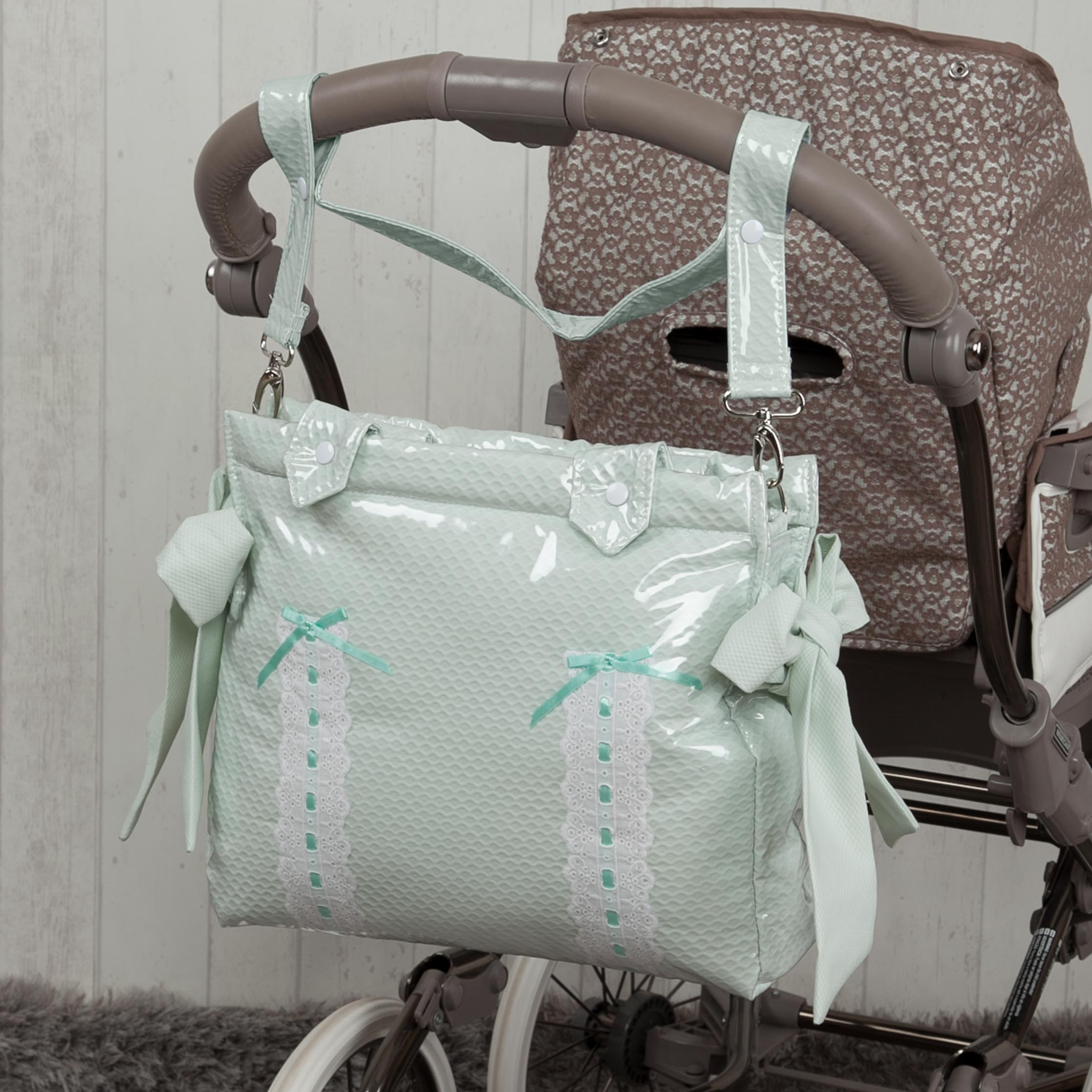 Un bolso de bebé verde menta con encaje blanco y lazos azules, colgado de un cochecito. Fondo beige.