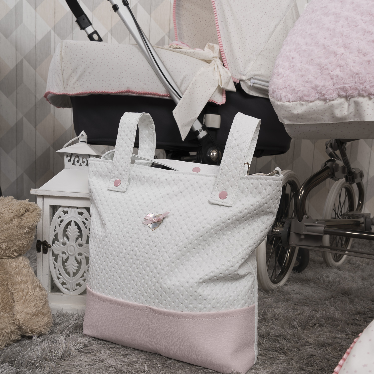 Bolso de bebé blanco con detalles rosas. Un oso de peluche y un cochecito en el fondo. Alfombra gris.