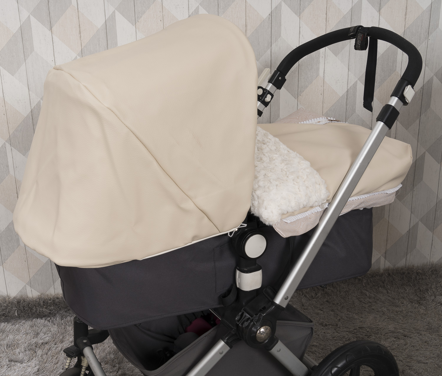 Cochecito de bebé color beige con capota crema y estructura gris oscuro, sobre alfombra gris.