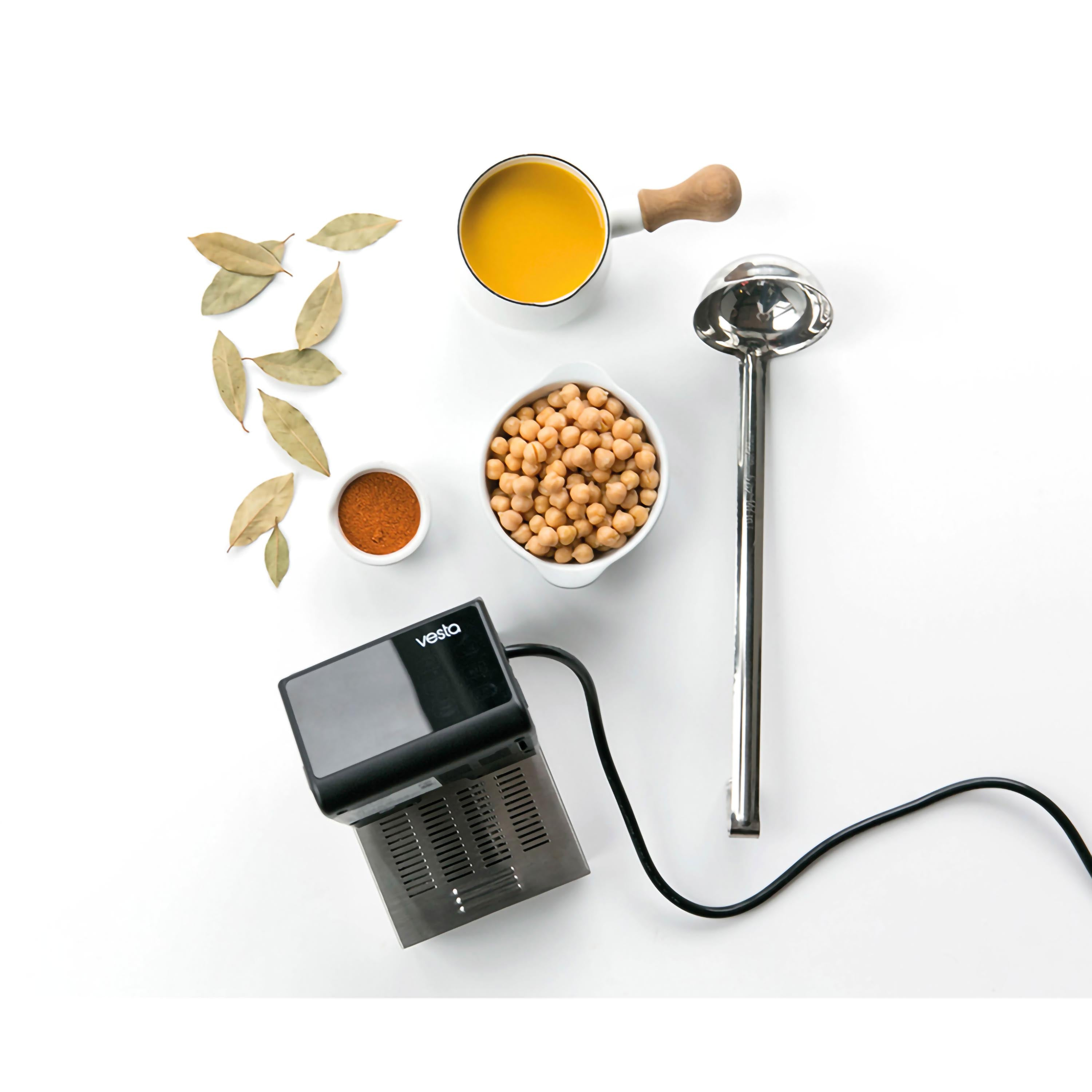 Vista dall'alto di ingredienti di cucina: ceci, spezie, un mestolo e un sous vide su una superficie bianca.