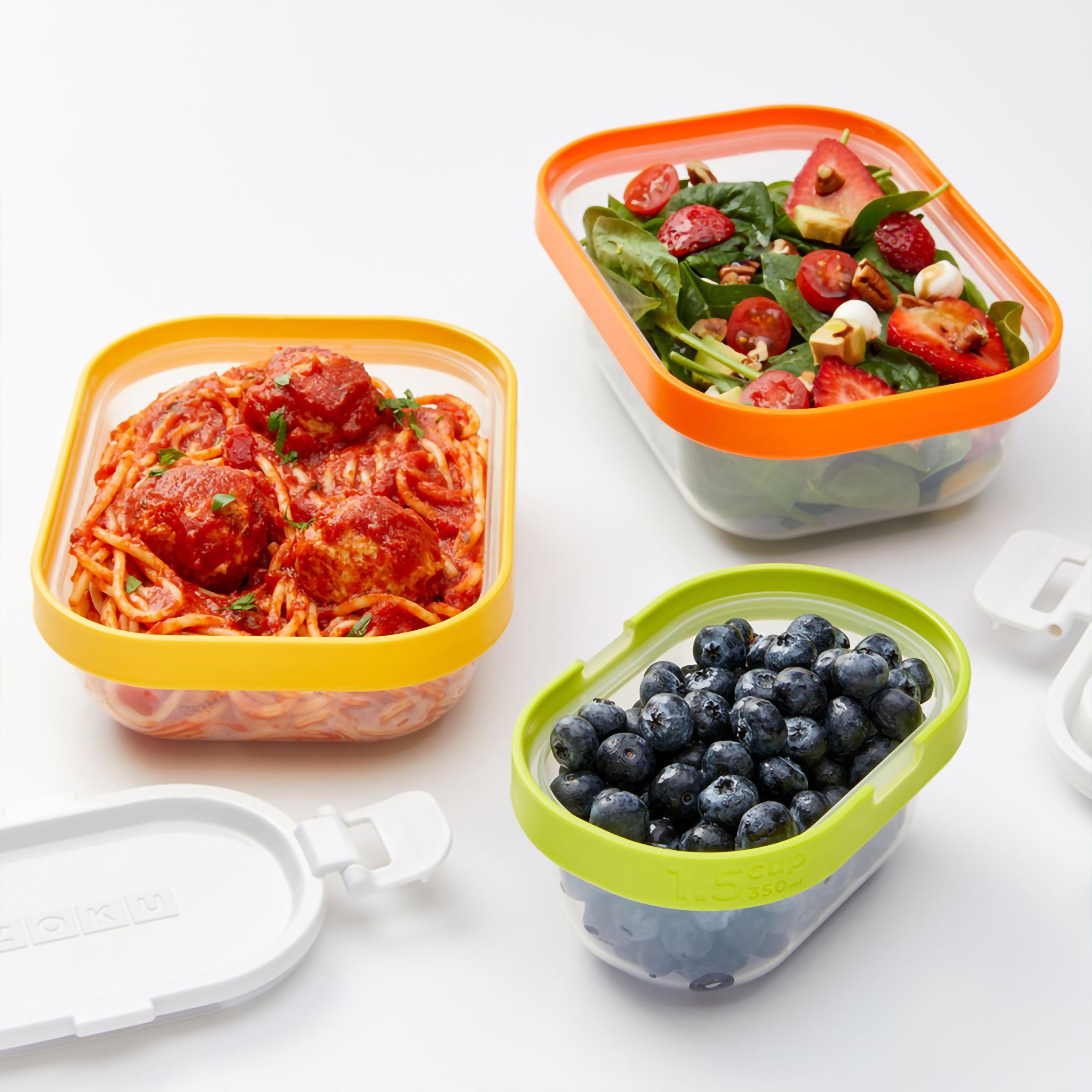 Drei quadratische, durchsichtige Lebensmittelbehälter mit farbigen Rändern, gefüllt mit Spaghetti, Salat und Blaubeeren, auf Weiß.