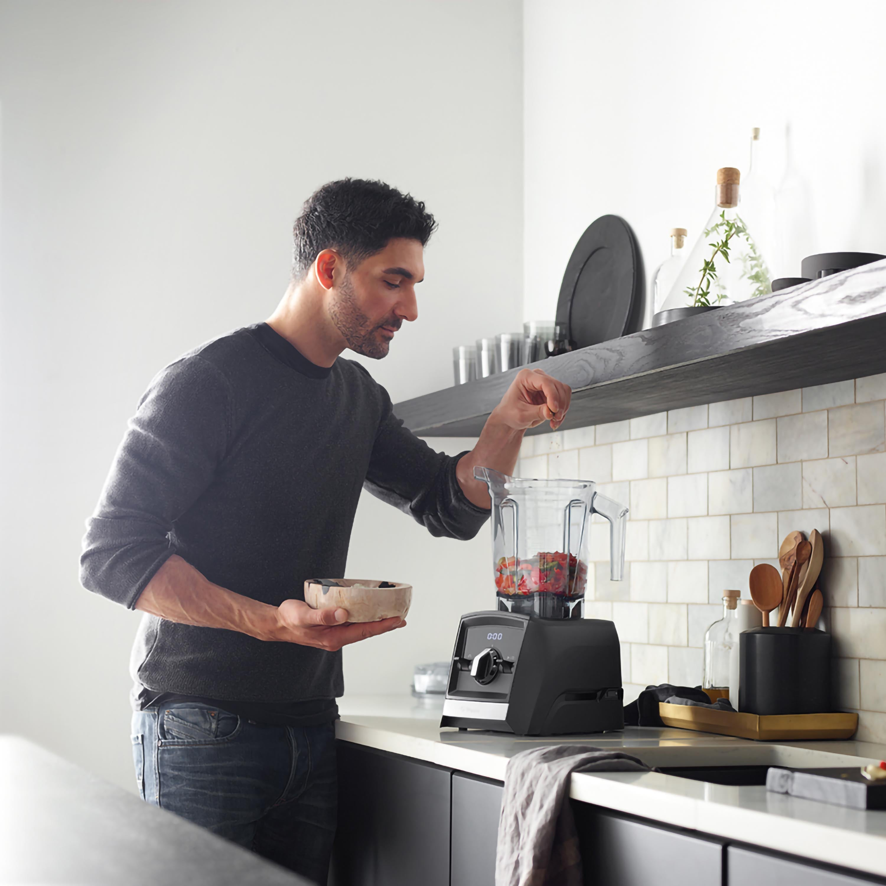 Uomo aggiunge ingredienti a un frullatore nero. Ciotola di cibo nella mano sinistra. Bancone della cucina.