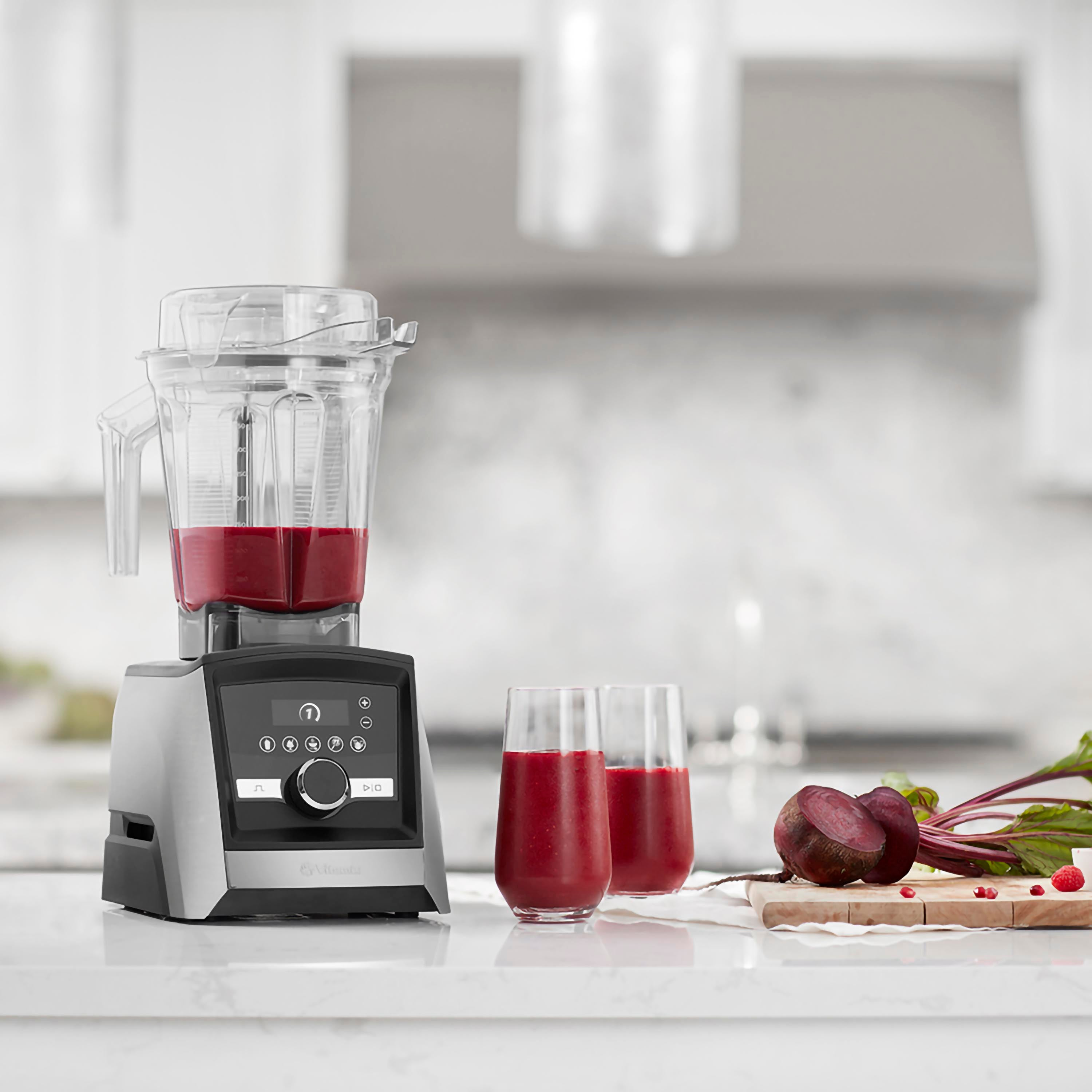 Mixer mit roter Flüssigkeit, zwei Gläsern Flüssigkeit und Rüben auf einem Holzbrett auf einer weißen Arbeitsplatte.