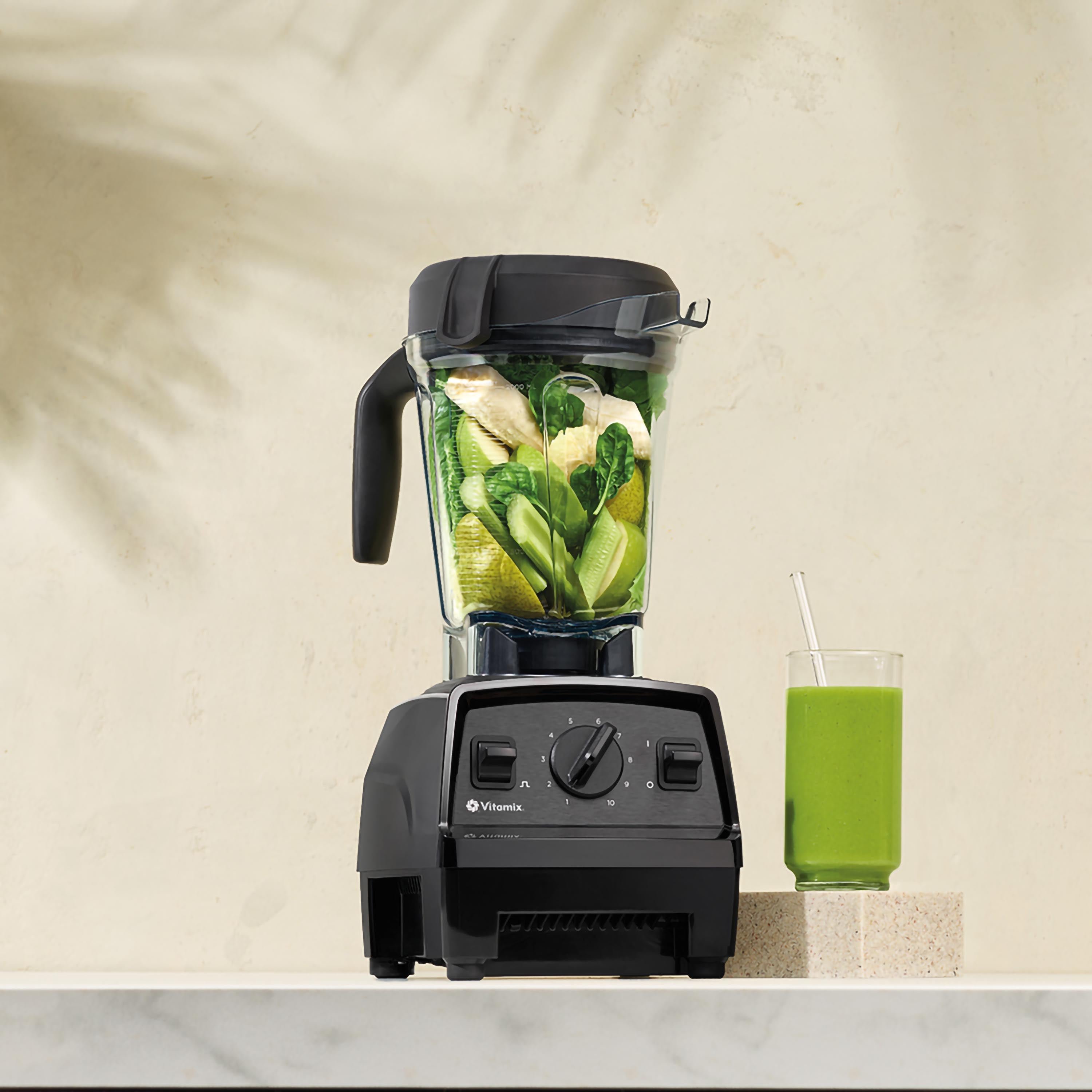 Ein schwarzer Mixer mit grünen Zutaten und einem Glas grünen Smoothie.