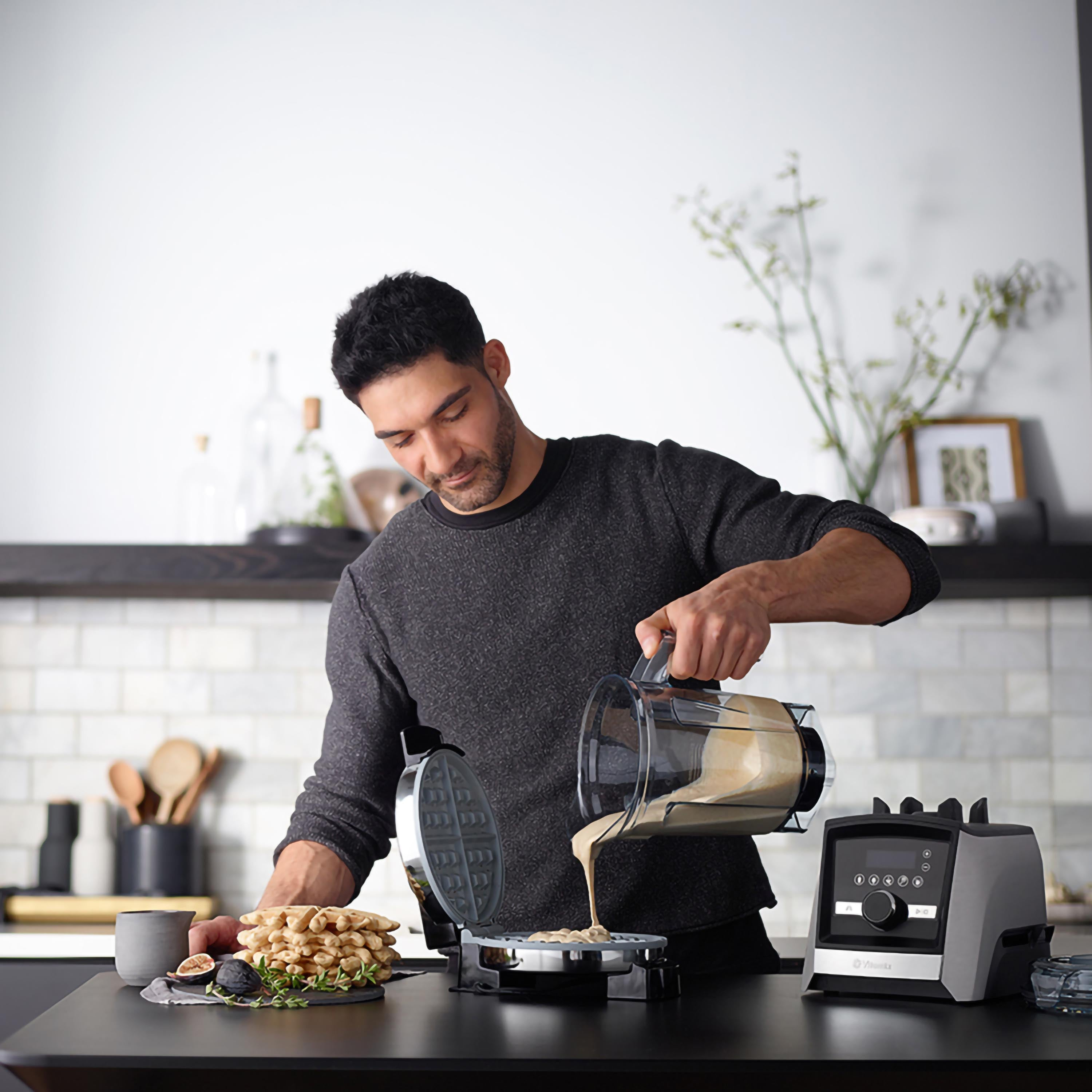 Mann gießt Teig in ein Waffeleisen, neben einem Mixer und Waffeln.