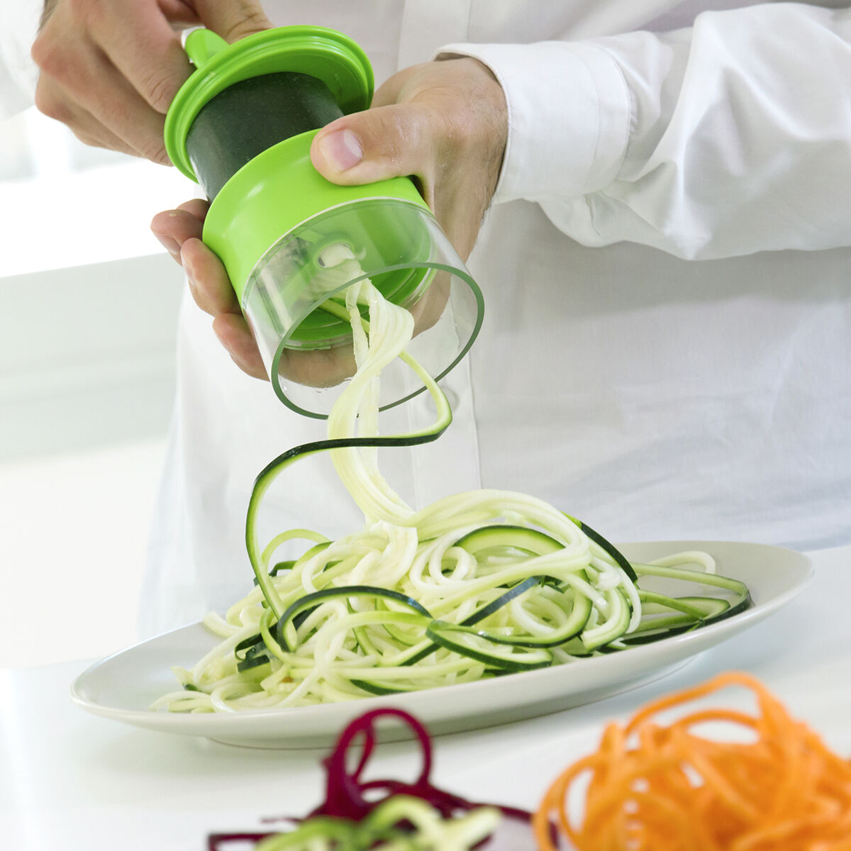 Mano che usa uno spiralizzatore verde per creare spaghetti di zucchine. Gli spaghetti cadono in un piatto bianco.