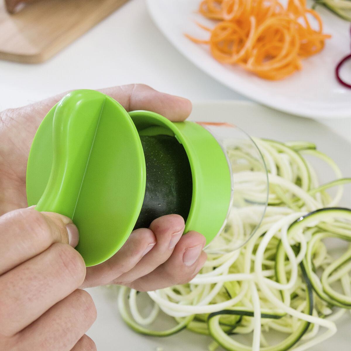 Mani che tengono uno spiralizzatore verde con zucchine. Spaghetti di zucchine e carote accanto.