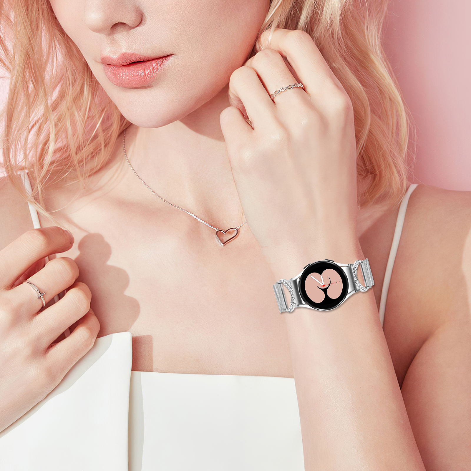 Il polso di una donna con un orologio d'argento, una collana a cuore e anelli. Sfondo rosa.
