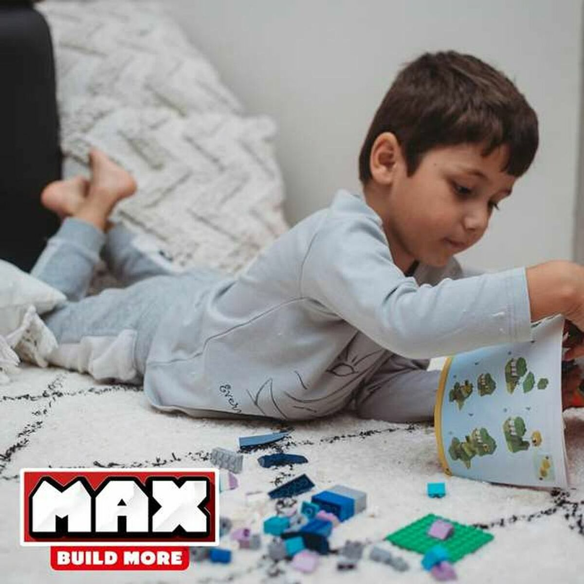 Junge, der auf einem Teppich liegt, mit LEGO-Steinen spielt und ein Buch liest, MAX-Logo sichtbar.