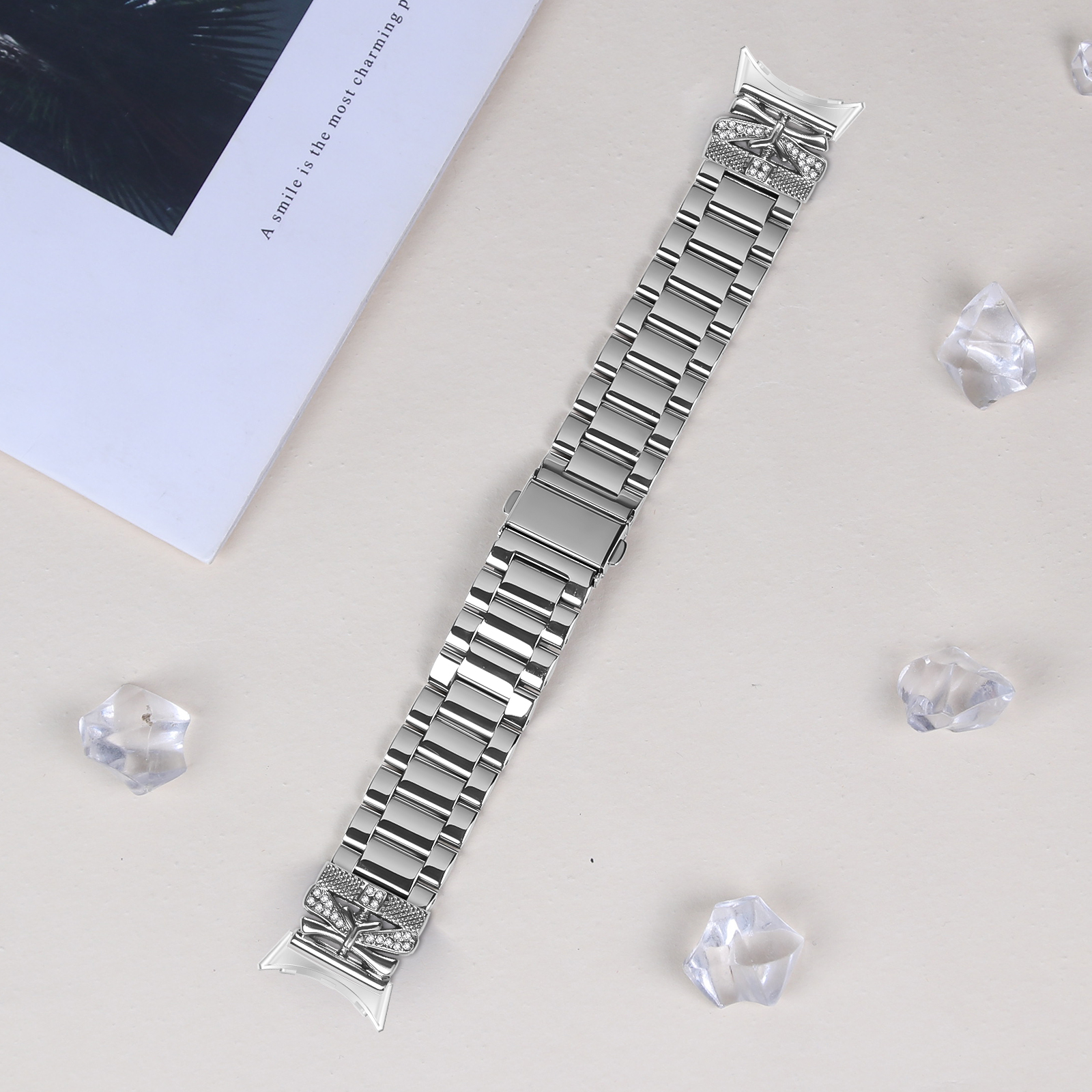 Silbernes Uhrenarmband mit dekorativen Elementen und klaren Kristallen. Ein weißes Papier und Kristalle sind drum herum.