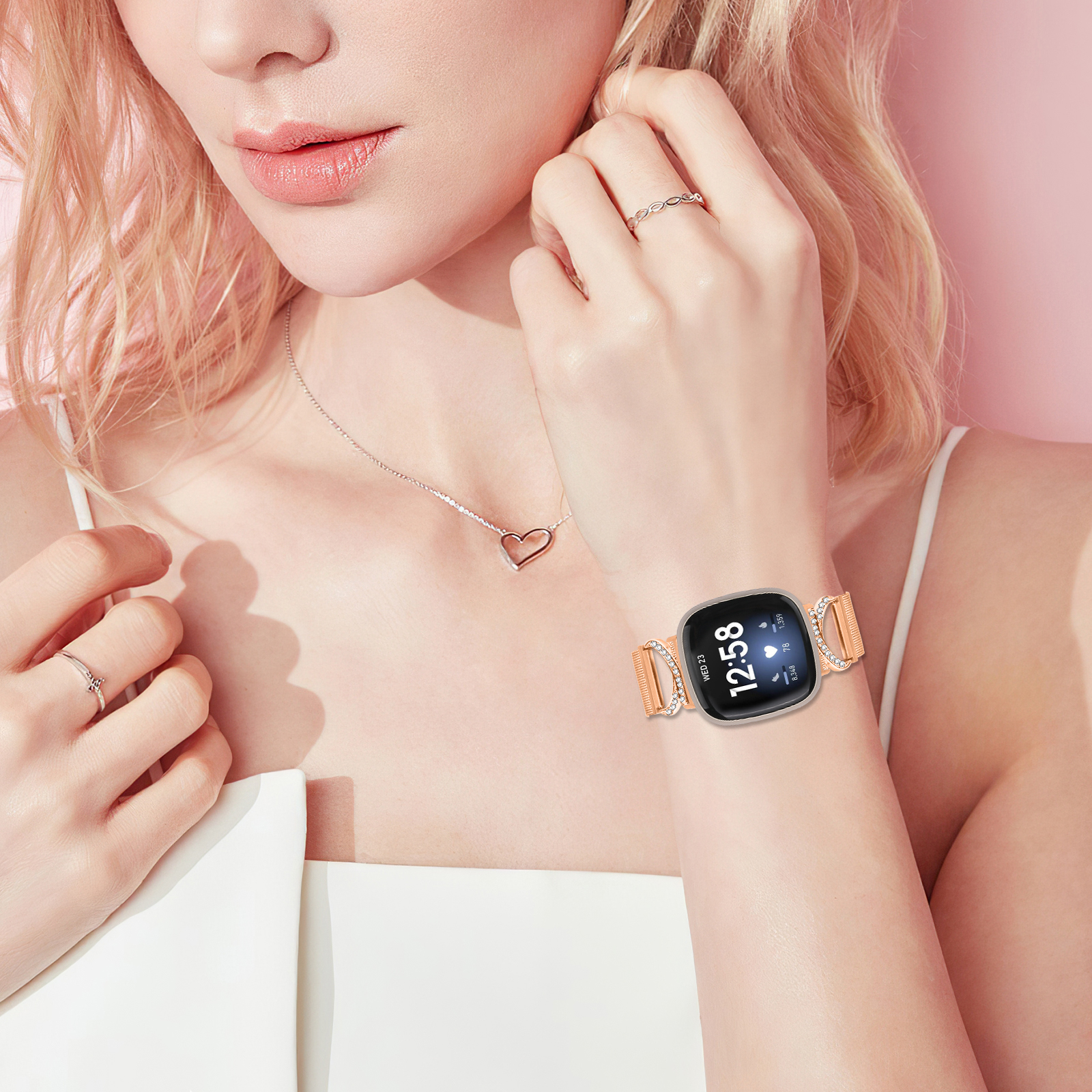 Handgelenk einer Frau mit Smartwatch, Ringen und Halskette vor rosa Hintergrund.
