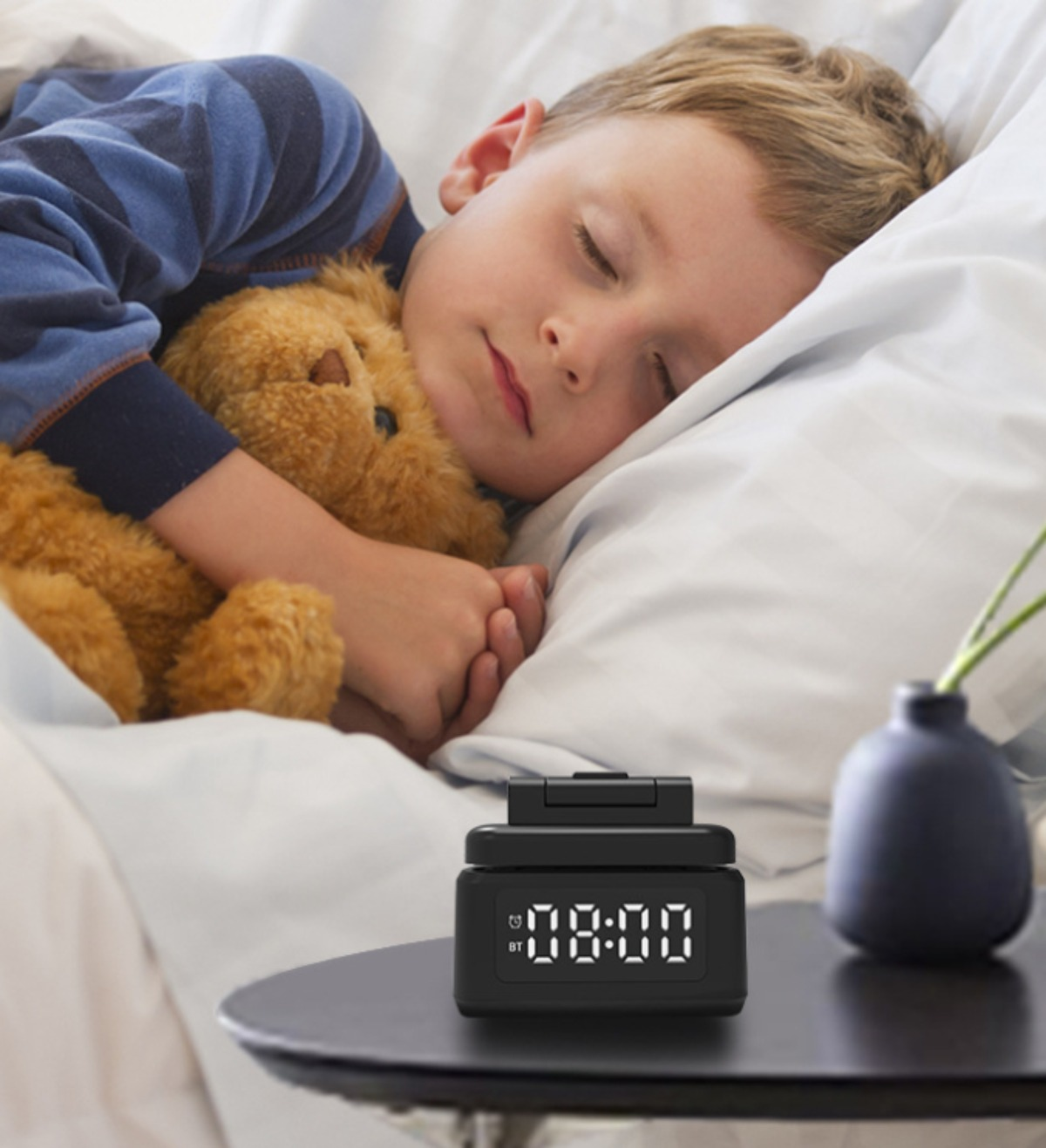 Ein schlafendes Kind im Bett mit einem Teddybären und einem digitalen Wecker auf dem Nachttisch.