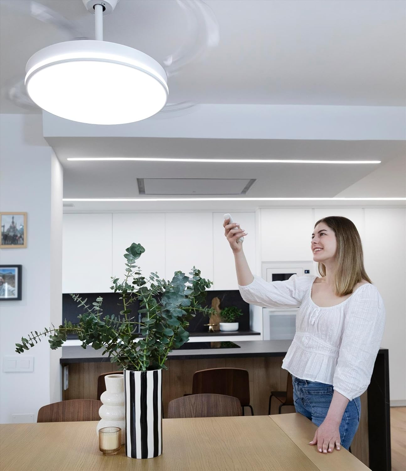Mujer controlando un ventilador de techo con un mando. Florero con plantas en la mesa.