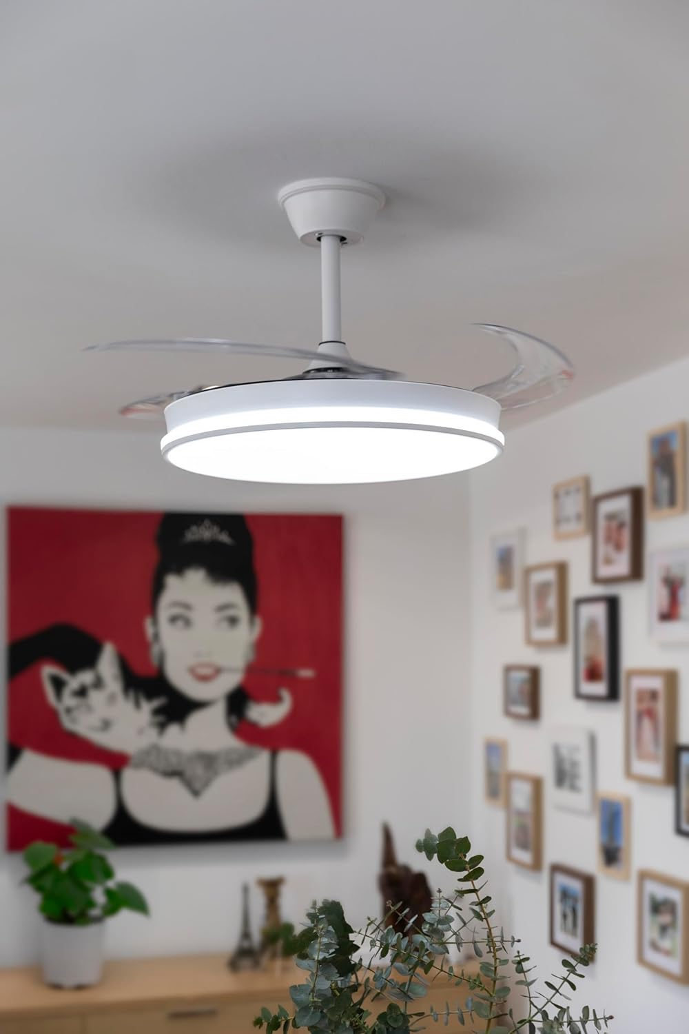 Ventilador de techo blanco con luz. Pintura de mujer en el fondo. Plantas en primer plano.