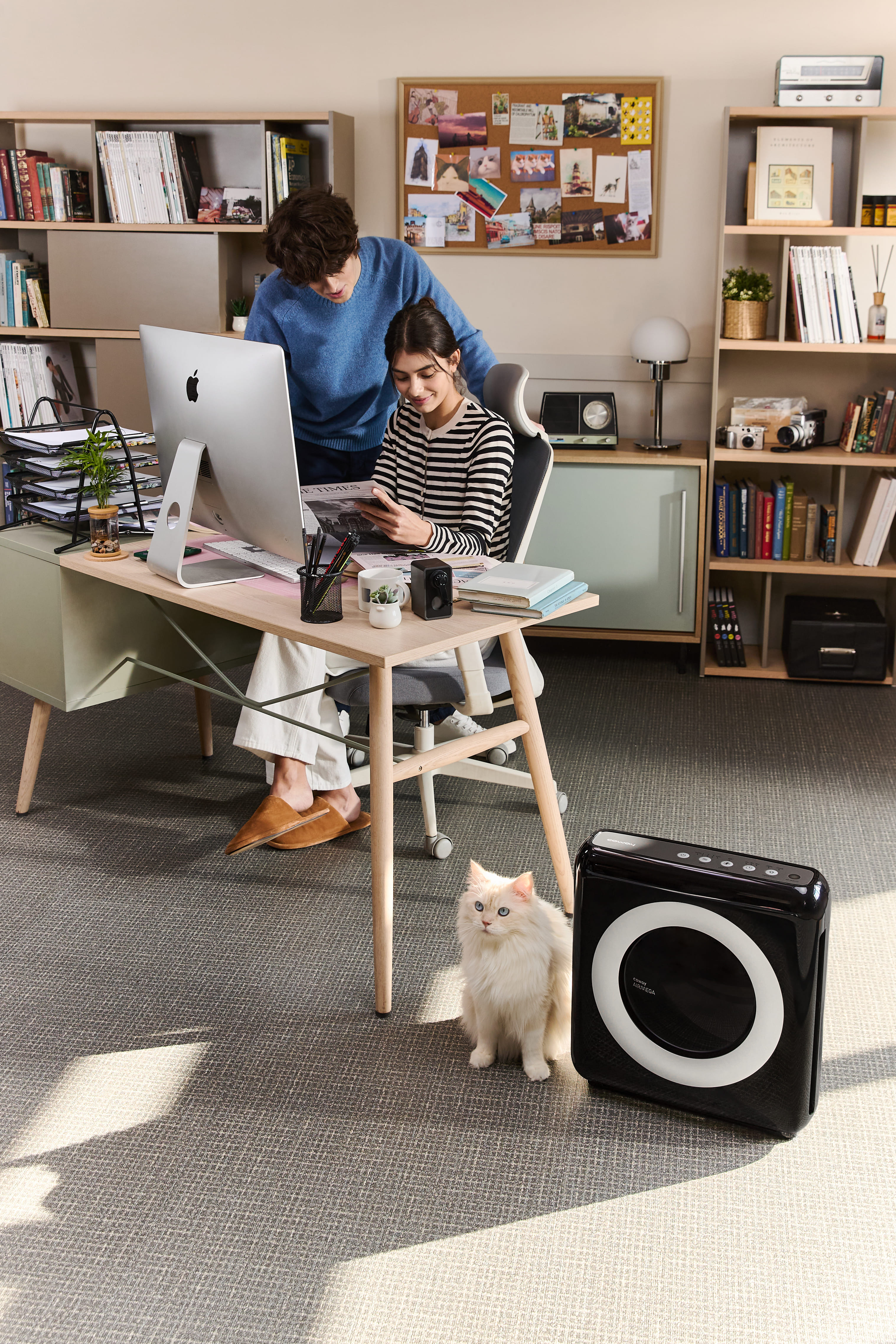 Zwei Personen in einem Büro mit Katze, Schreibtisch mit Computer, Büchern und Pflanzen, sowie Soundsystem.
