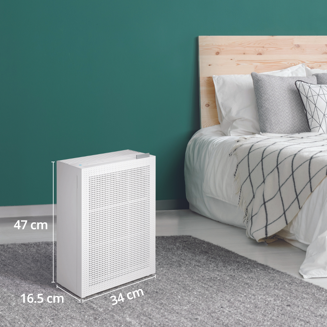 Ein weißer Luftreiniger steht neben einem Bett in einem Raum mit teal Wand. Maße sind enthalten.