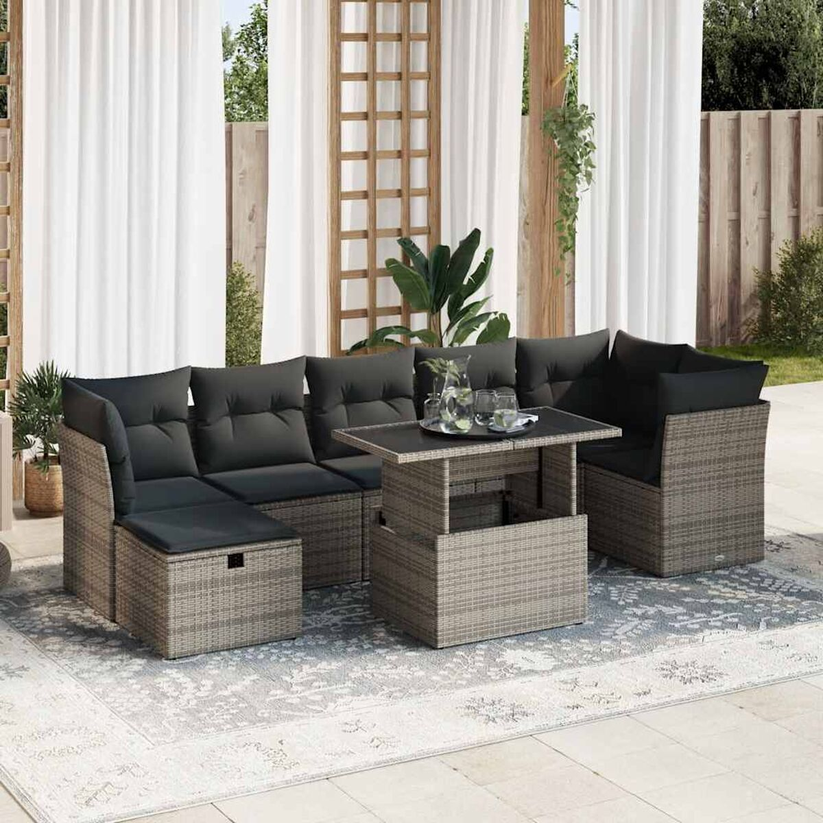 Gartenmöbel-Set aus Weide mit schwarzen Kissen und einem Tisch auf einem gemusterten Teppich. Vorhänge und Pflanzen sichtbar.