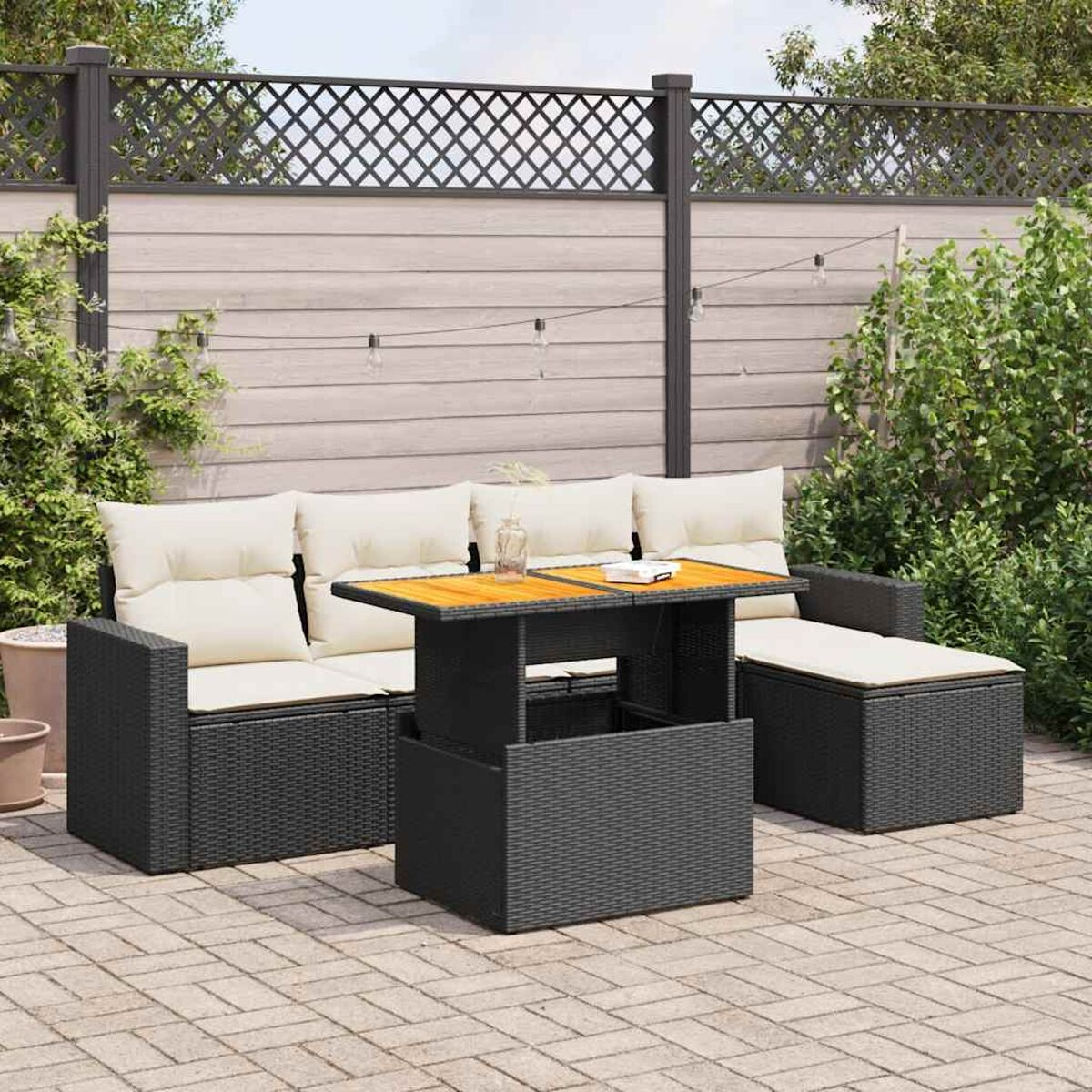 Divano in rattan nero da esterno con cuscini bianchi e un tavolo in legno, su un patio con recinzione e piante.