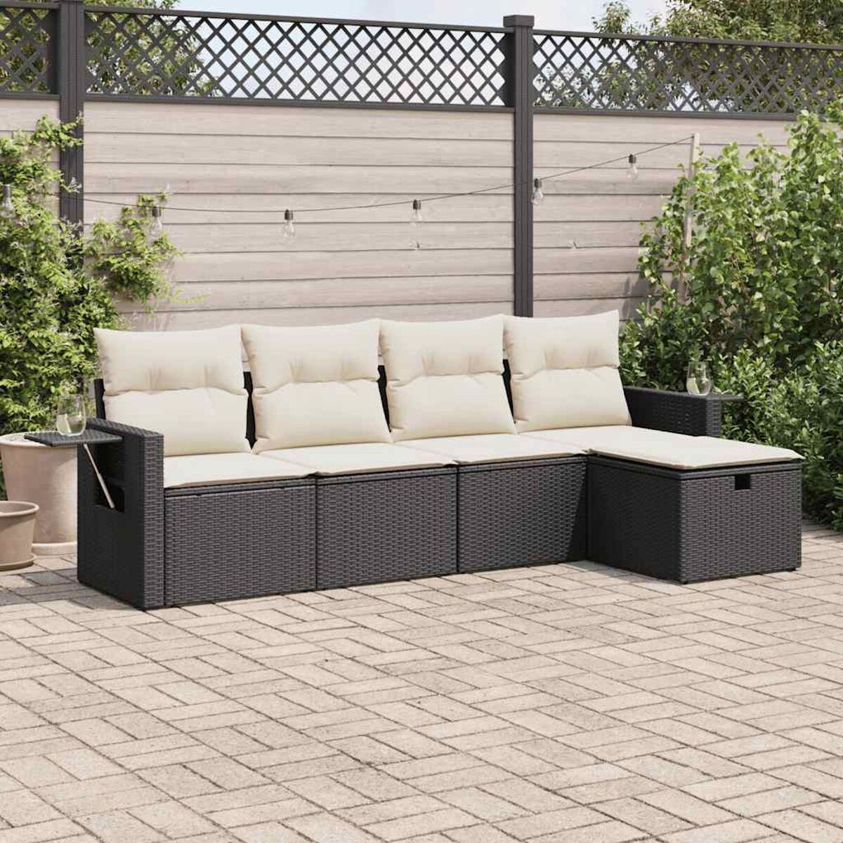 Dunkles Ecksofa aus Korbgeflecht mit Kissen auf einer Backstein-Terrasse. Zaun und Pflanzen im Hintergrund.