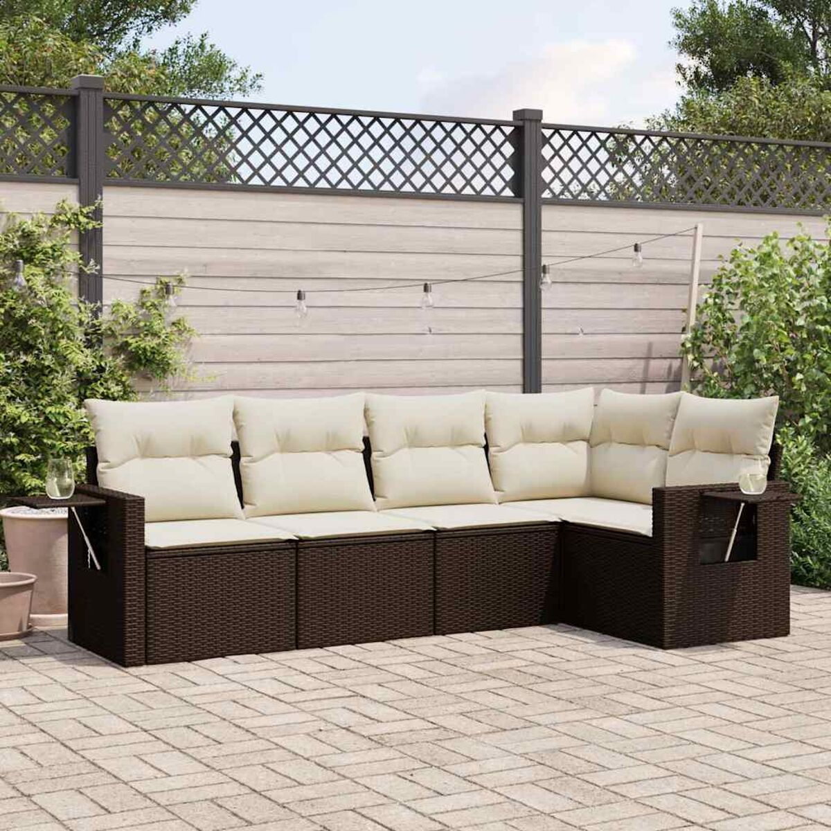 Braunes Korbsesselsofa mit cremefarbenen Kissen auf einer Ziegelterrasse, mit Zaun und Grün im Hintergrund.