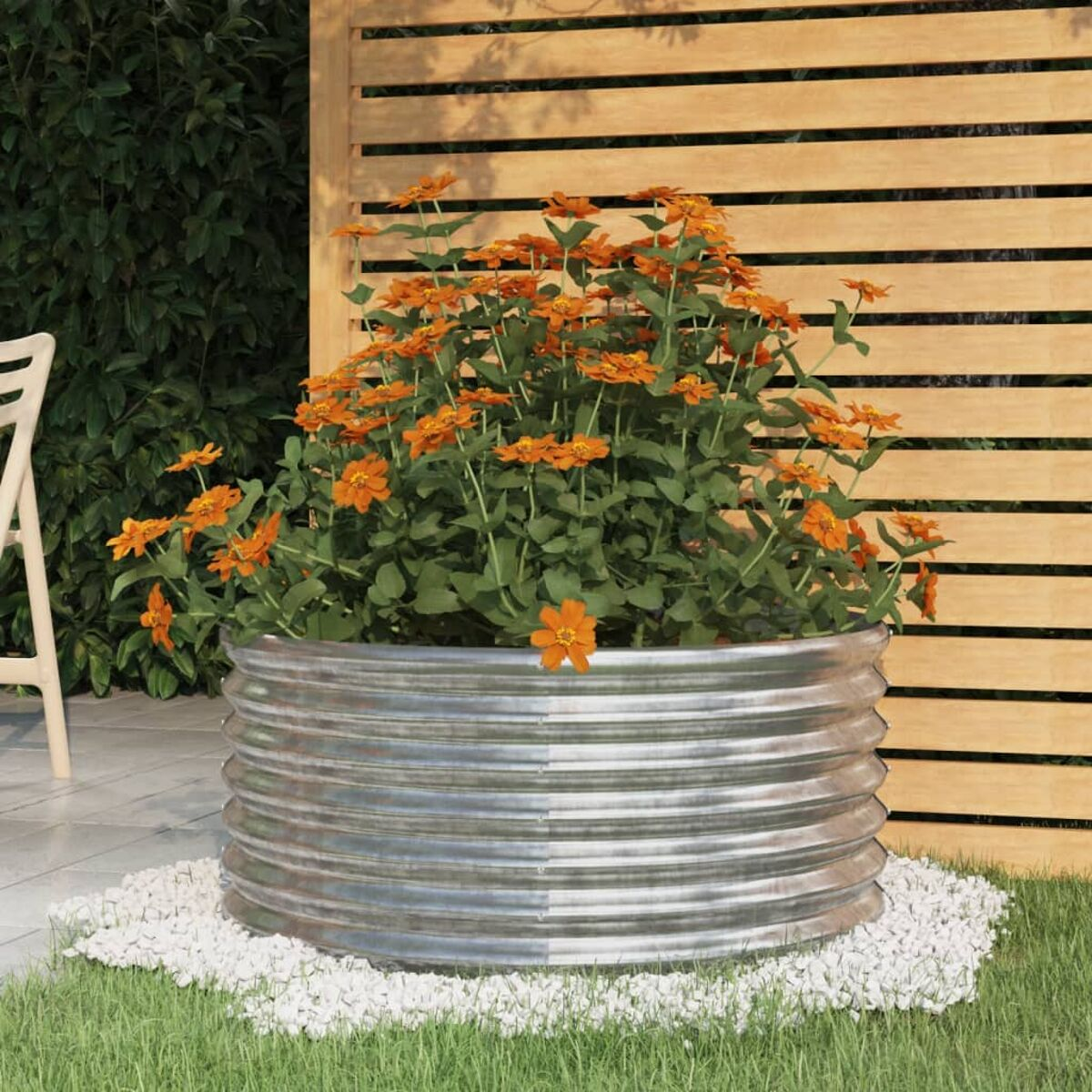 Metallpflanzer mit orangefarbenen Blumen. Weiße Steine, Holzzaun und grünes Gras.