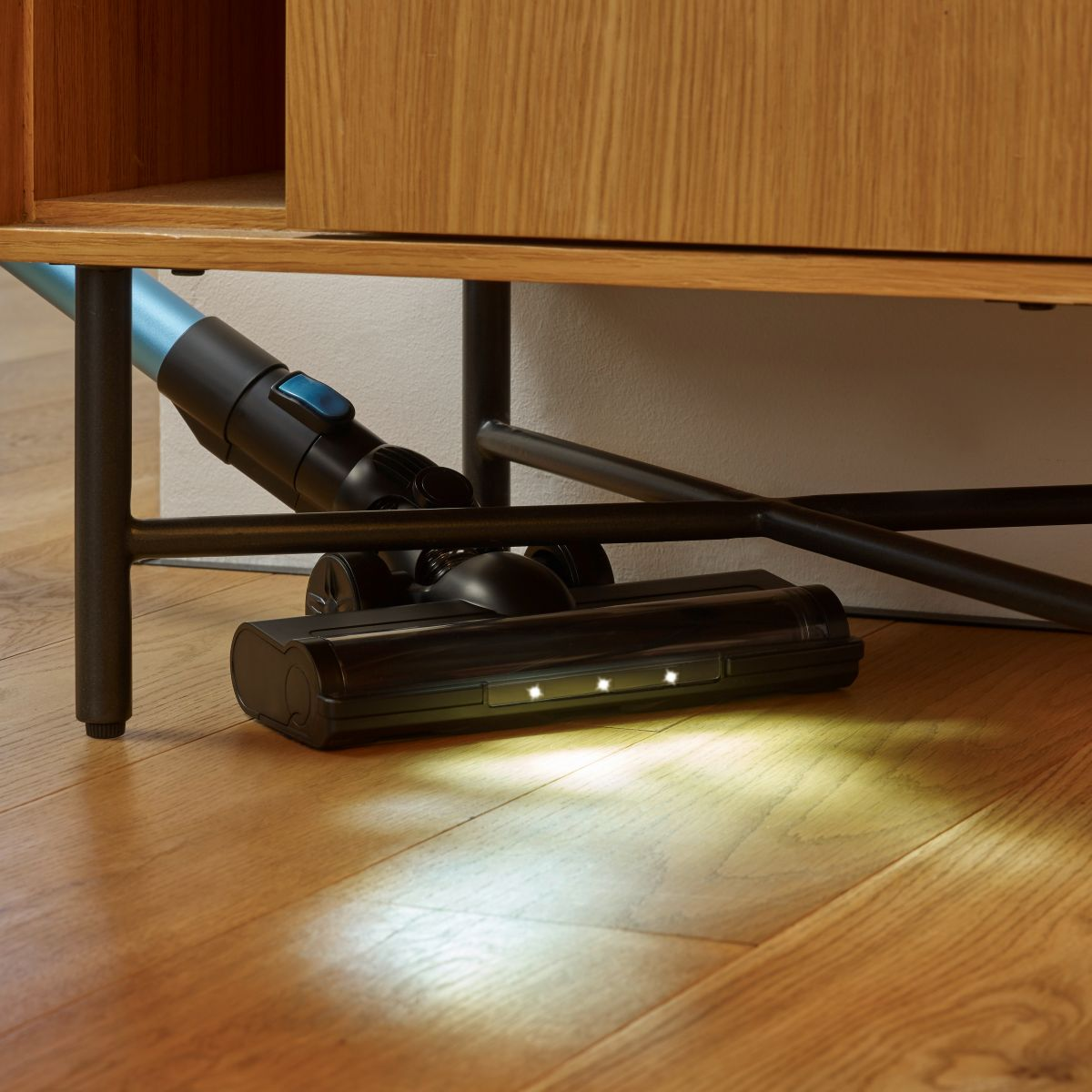 Cabezal de aspiradora negro bajo mueble de madera, en suelo de madera con luz brillante.