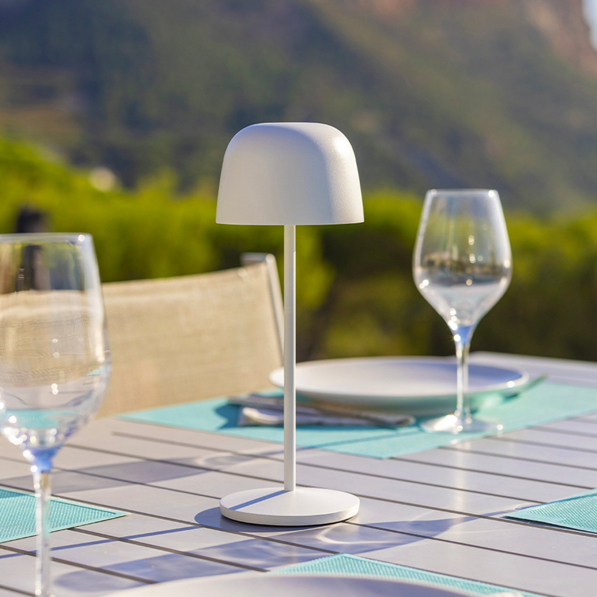 Weiße Lampe auf einem Tisch mit Weingläsern und Tellern. Grüne Bäume sind im Hintergrund.