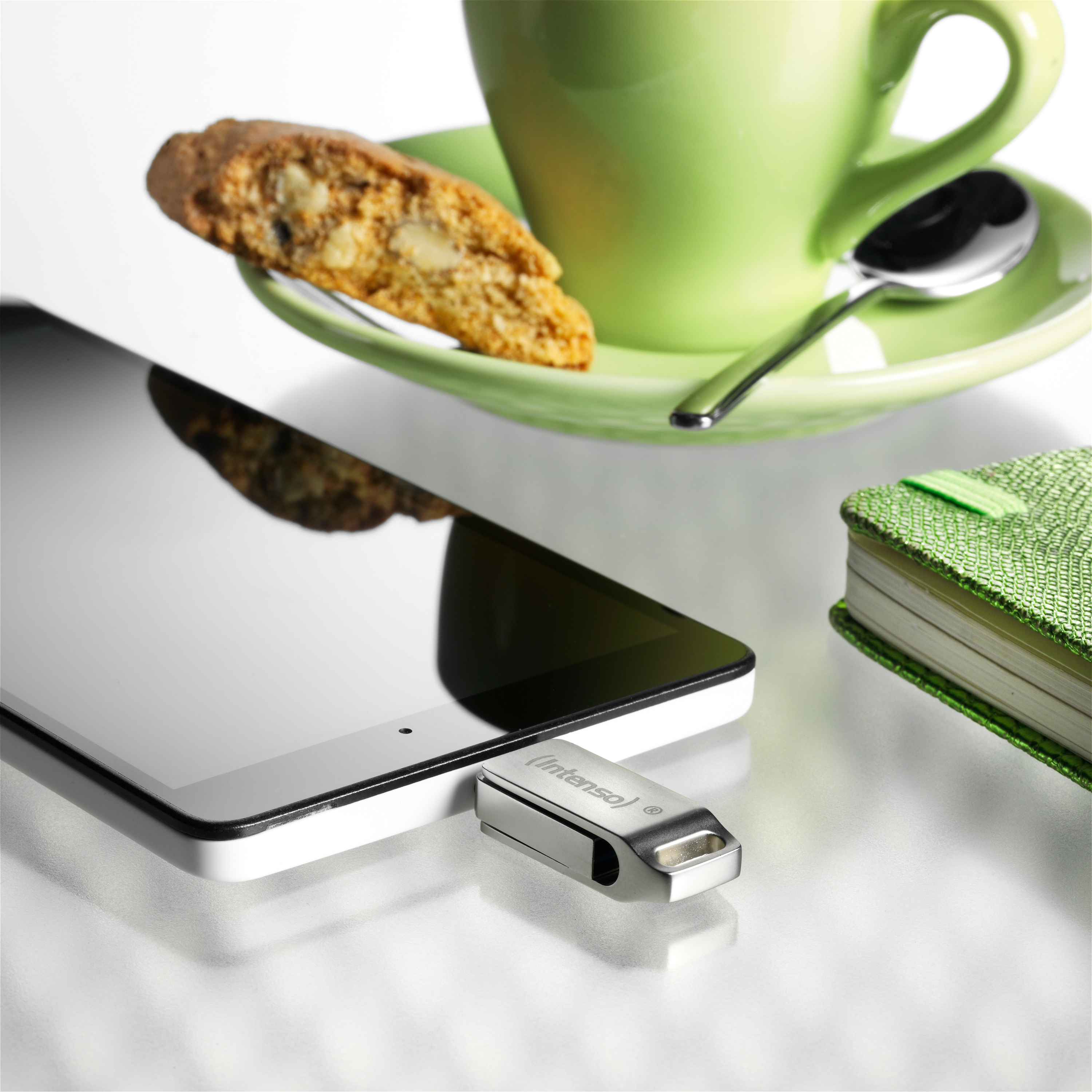 Tablet, biscotti, zielony kubek i spodek oraz srebrny pendrive z napisem Intensa.