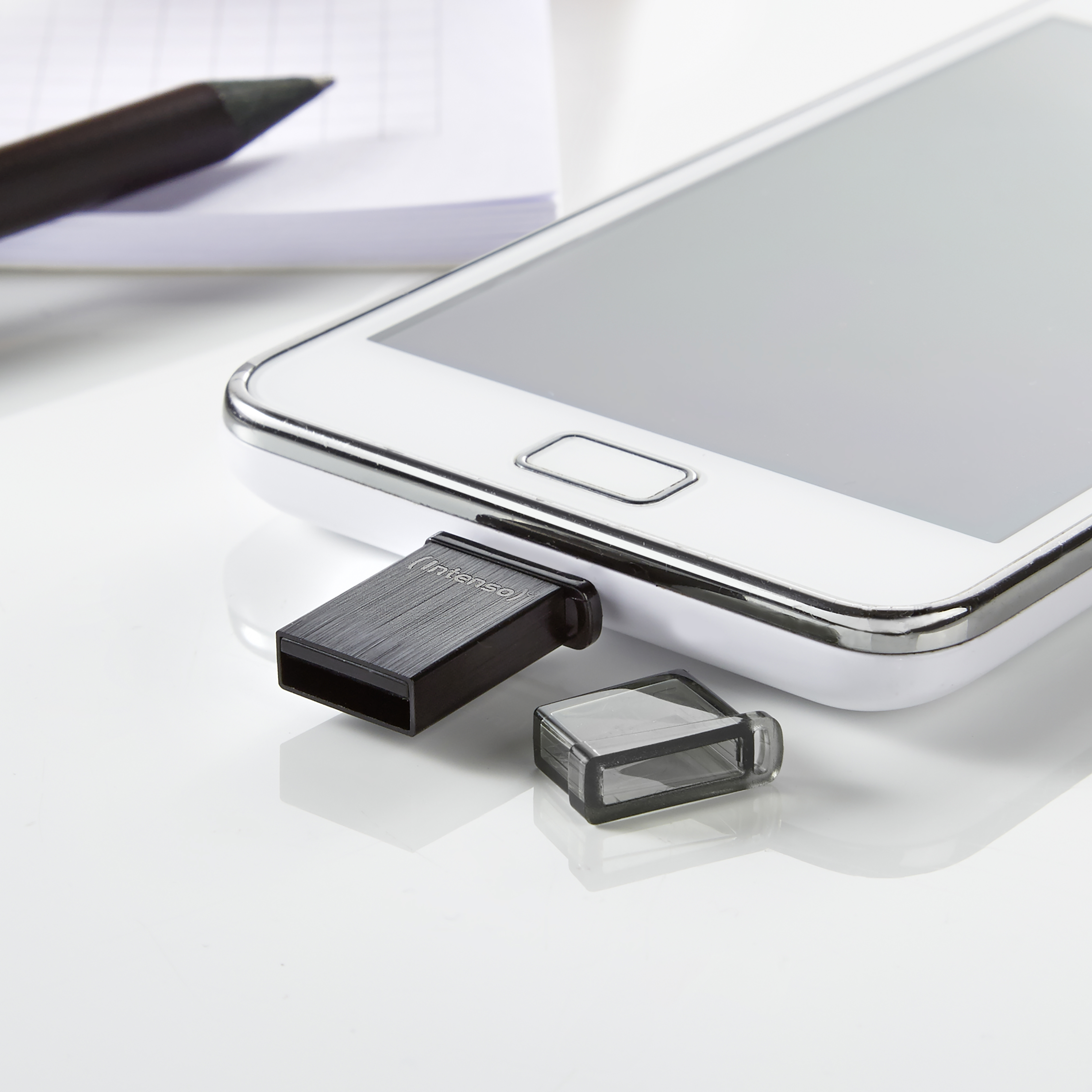 Smartfon podłączony do czarnego dysku USB. Obok napędu znajduje się nasadka. Tłem jest biała powierzchnia.