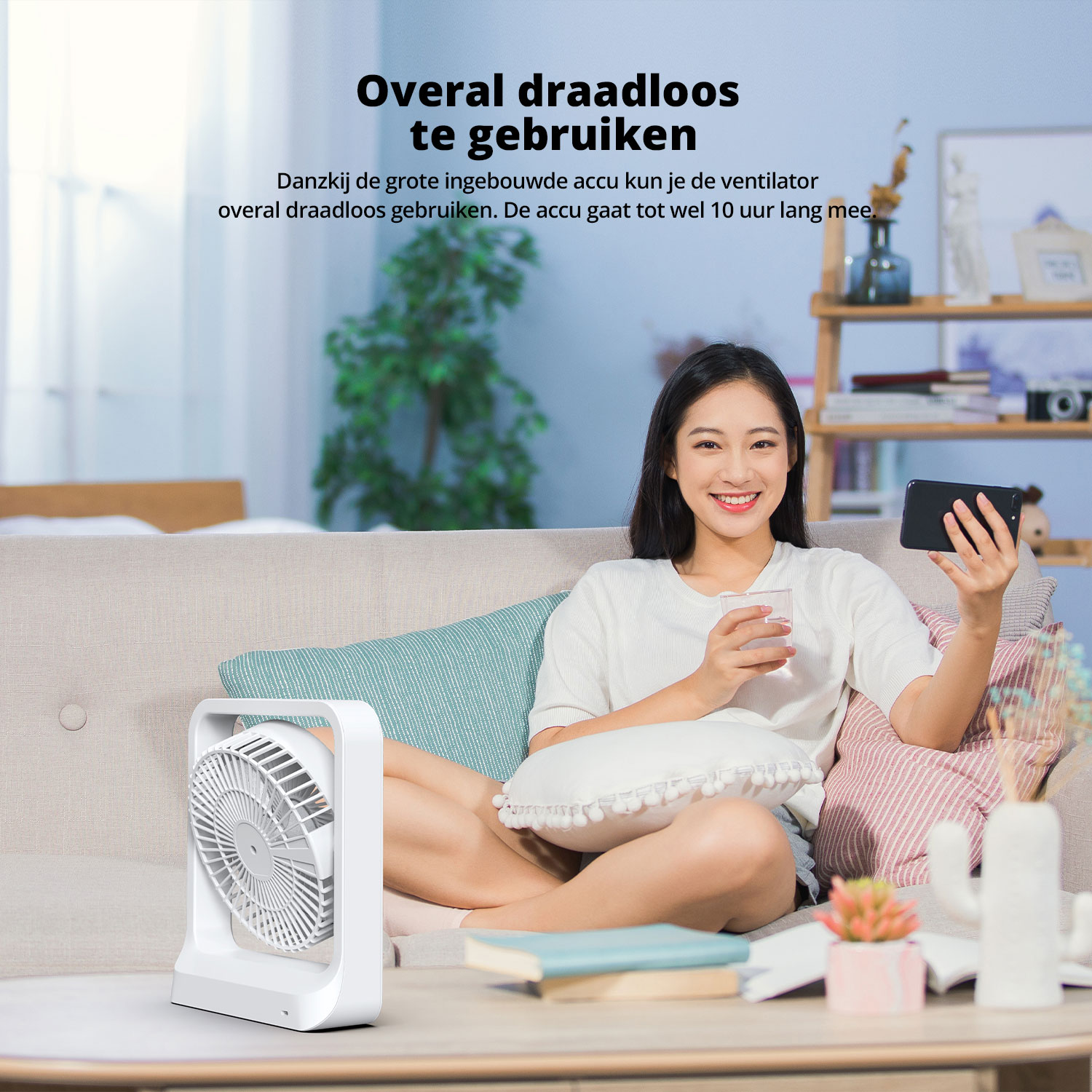 Femme tenant un téléphone et un verre assise sur un canapé. Un ventilateur est placé à gauche. Un cactus est en arrière-plan.